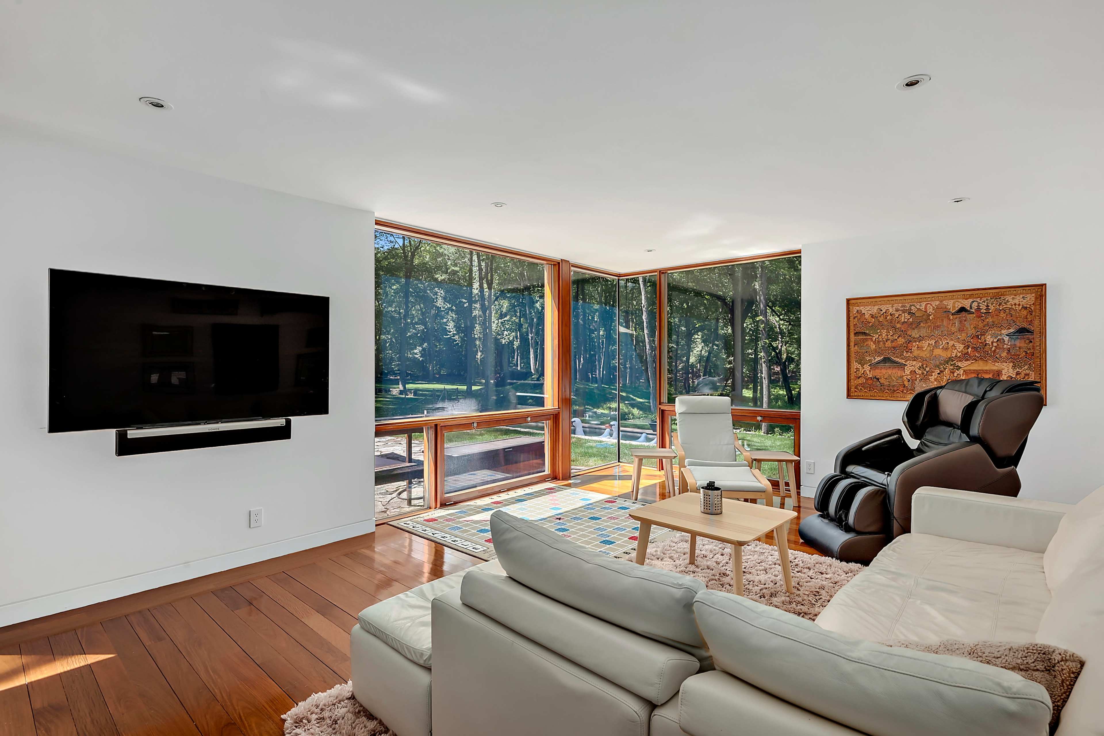 A modern living room features a television mounted on the wall, large windows with a view of a forest, and comfortable seating arranged around a small table.