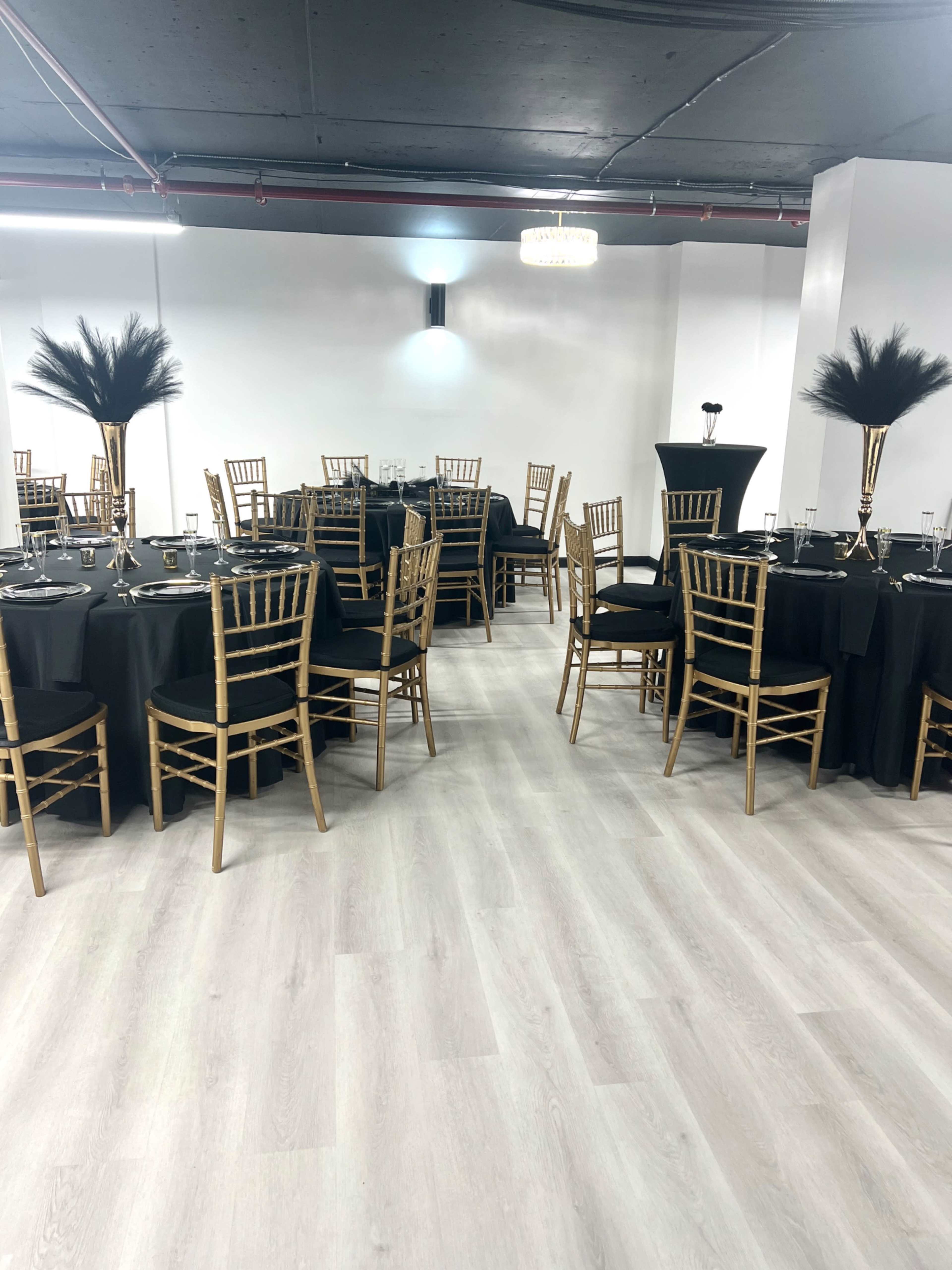 A well-arranged event space with round tables covered in black cloth, gold chiavari chairs, and centerpieces featuring tall, feather arrangements.