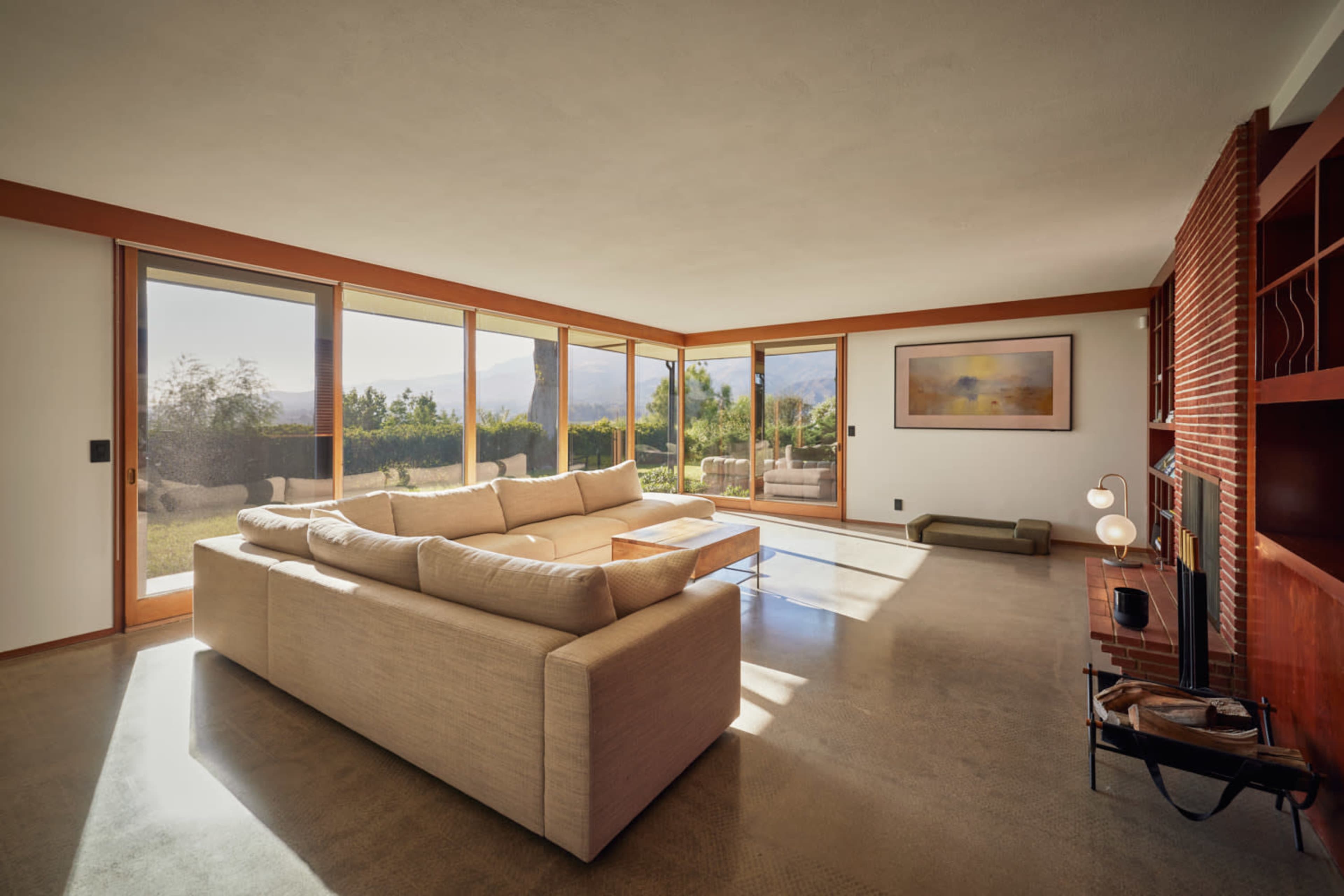The image shows a modern living room with large windows, a sectional sofa, a brick wall, and a view of an outdoor landscape.