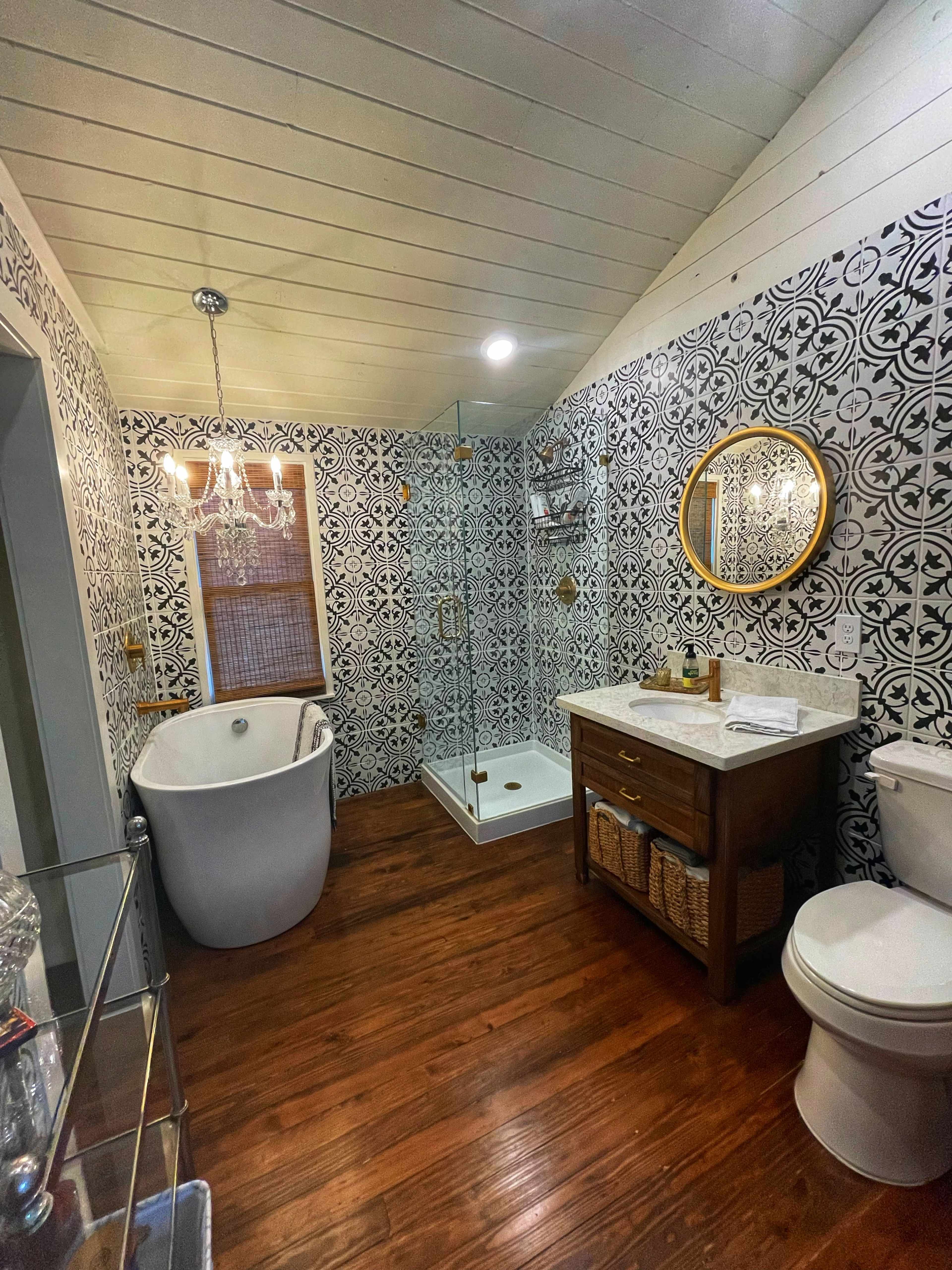The bathroom features a freestanding bathtub, a glass shower enclosure, a marble vanity with a round mirror, and patterned tile walls over hardwood flooring.