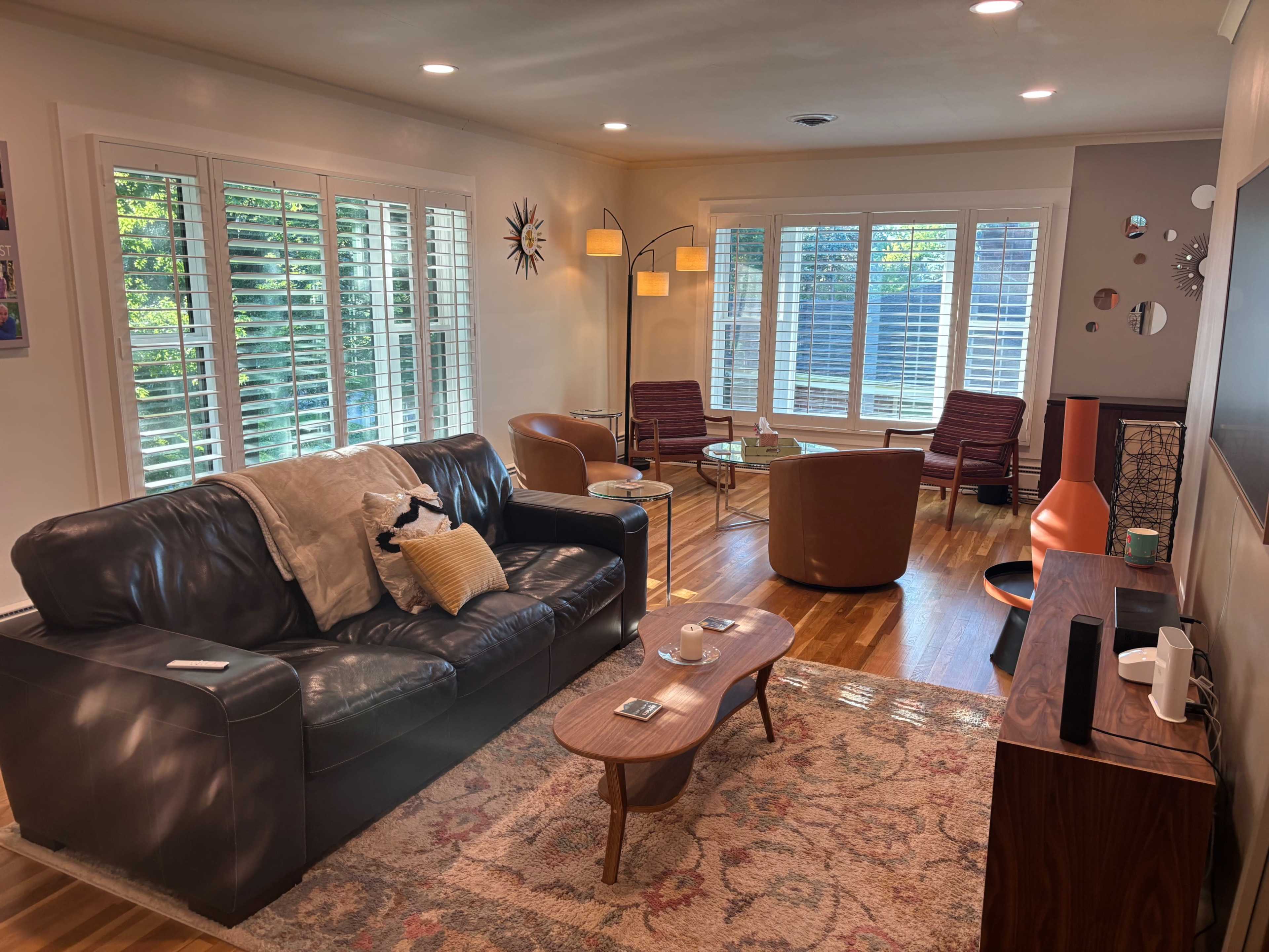 A light-filled living room features a leather sofa, two armchairs, a coffee table, and large windows with white shutters.