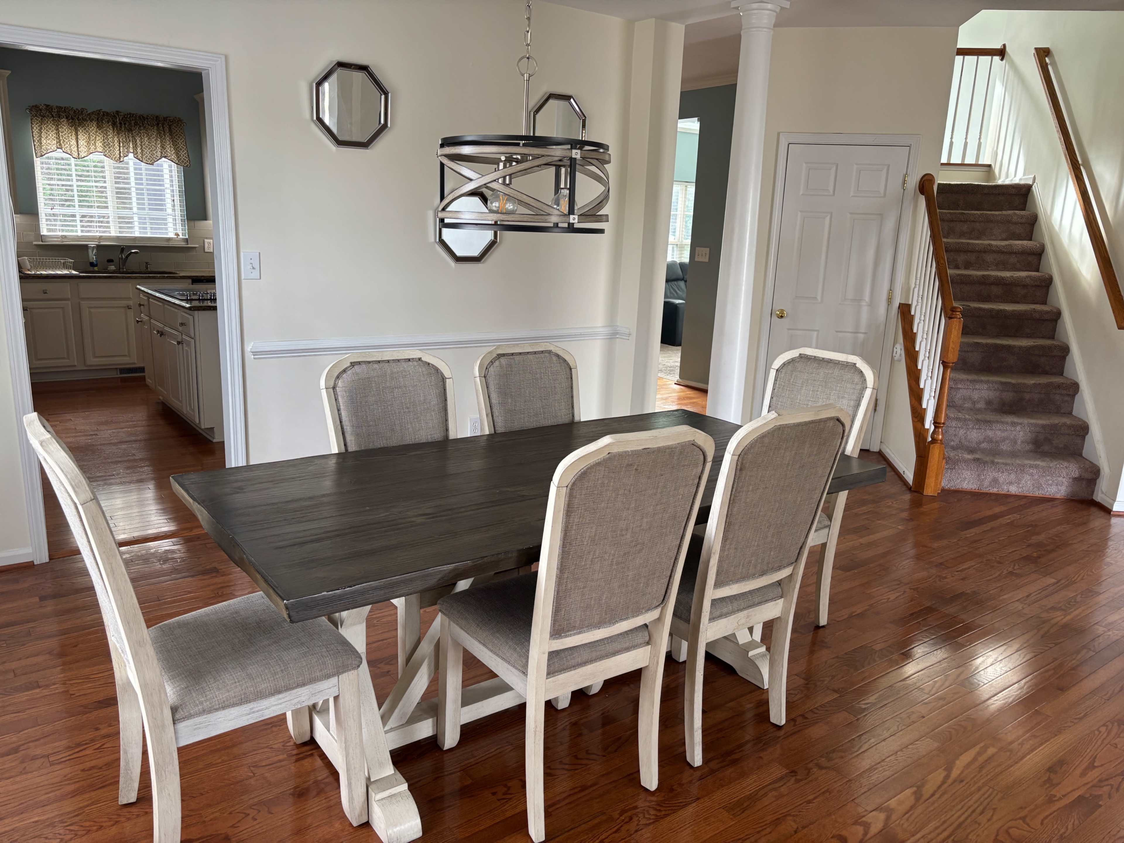 A dining room features a wooden table with six chairs, a modern light fixture above, and a staircase leading to an upper level.
