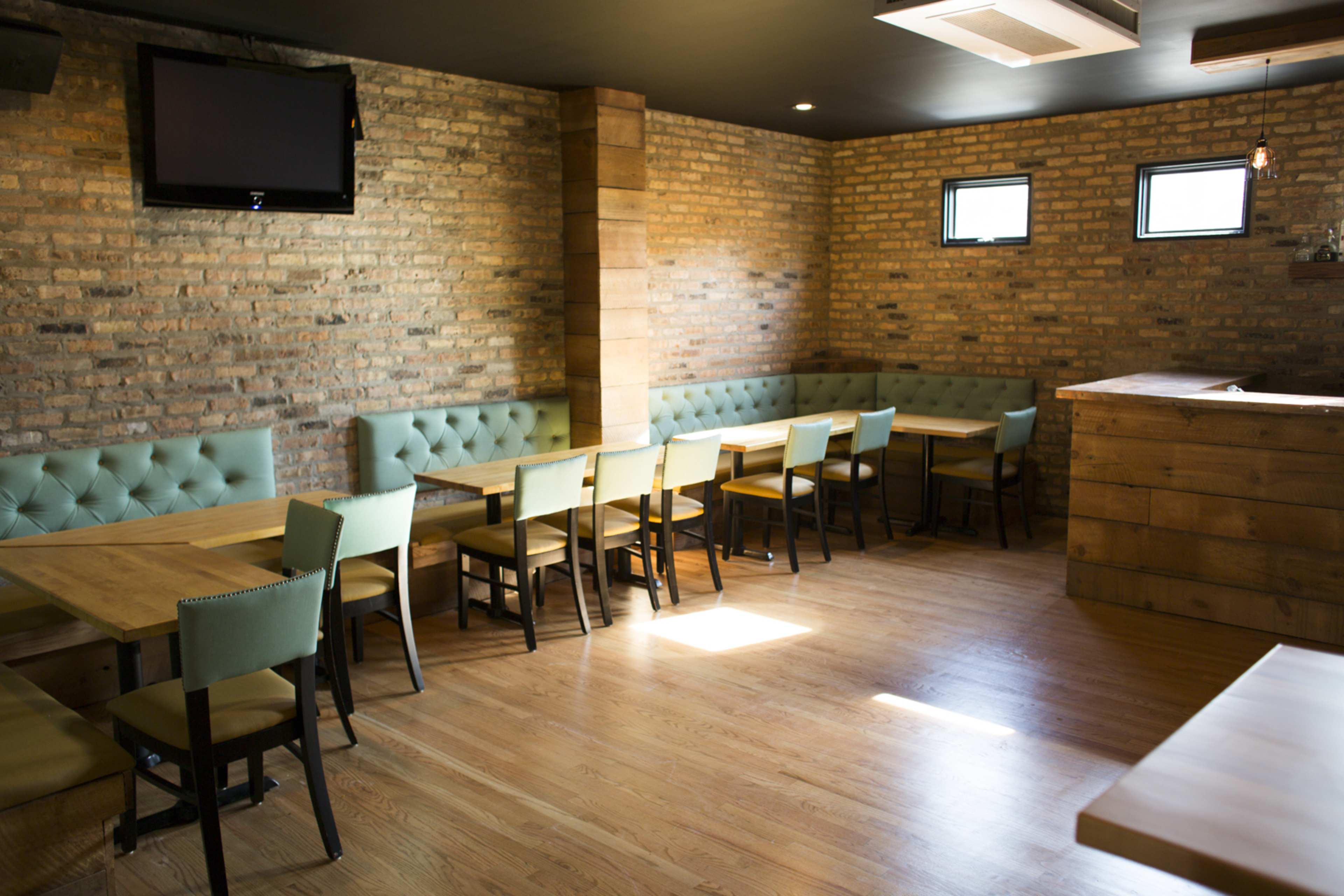 The image shows a restaurant interior with wooden flooring, brick walls, green upholstered seating, and several tables arranged for dining.