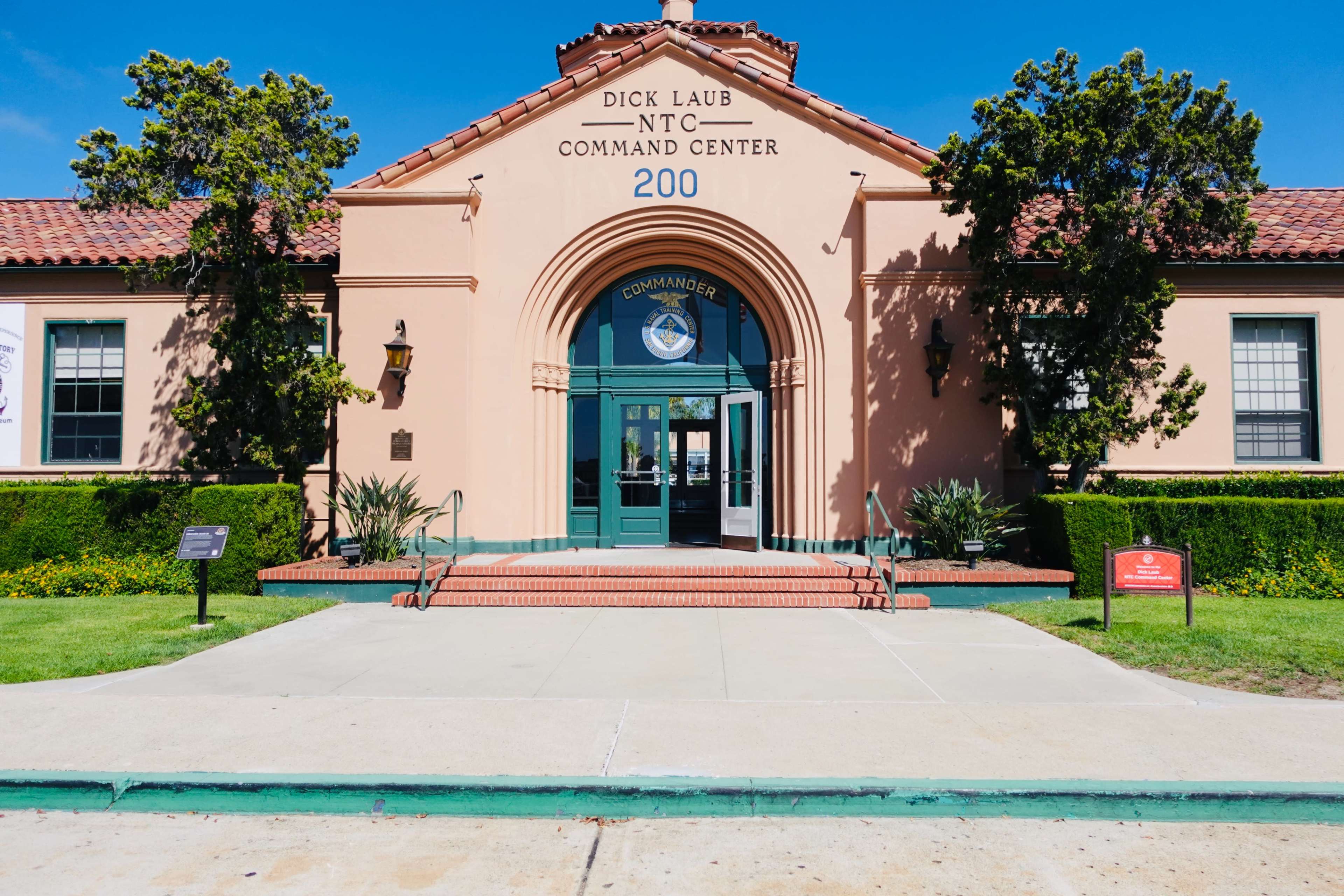 The image shows the entrance of the Dick Laub NT Command Center, featuring a terracotta roof, green doors, and landscaped greenery in front.