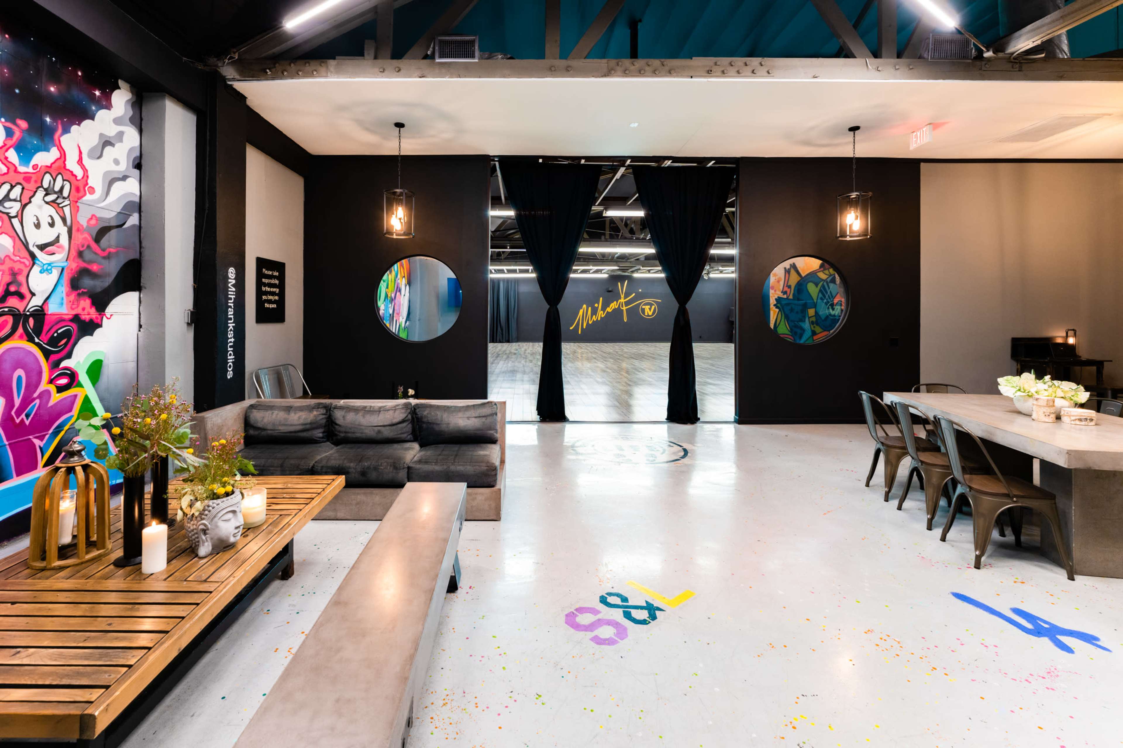 The image shows a contemporary studio space featuring a seating area with a sofa, a dining table, and colorful graffiti on the walls, leading to a large, open area beyond.