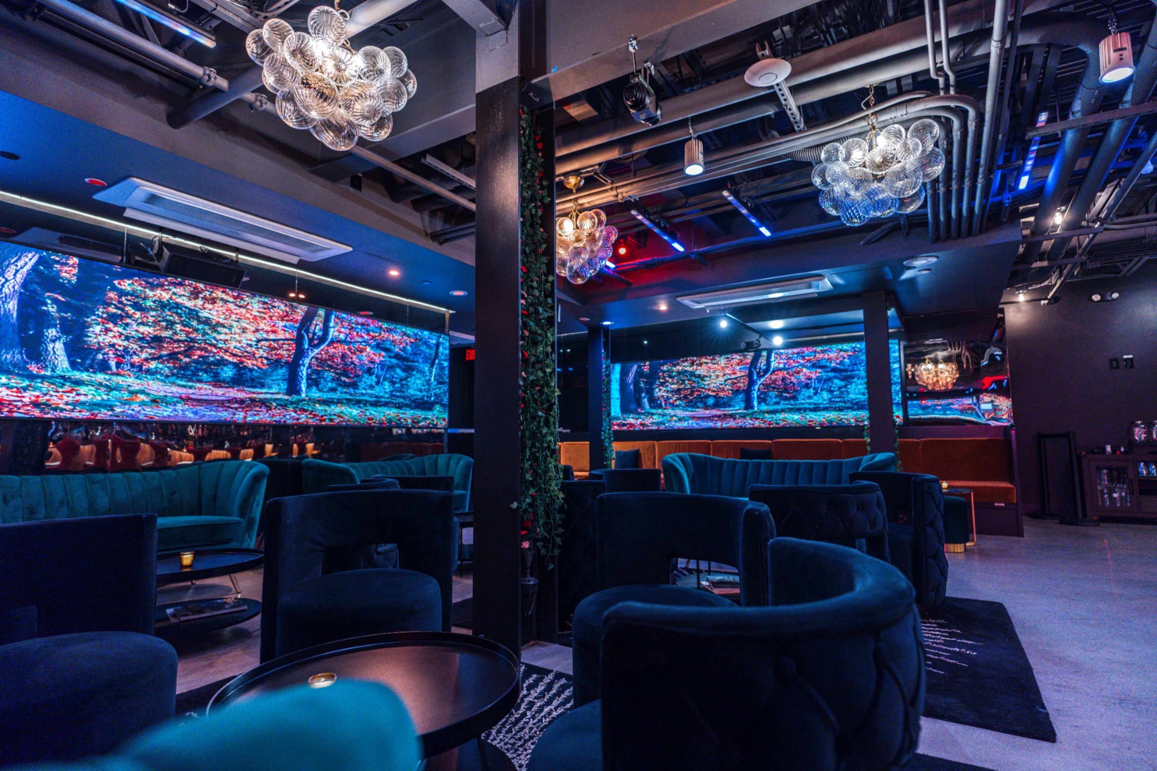 The image shows a modern lounge interior featuring blue velvet seating, decorative chandeliers, and large screens displaying colorful nature scenes on the walls.