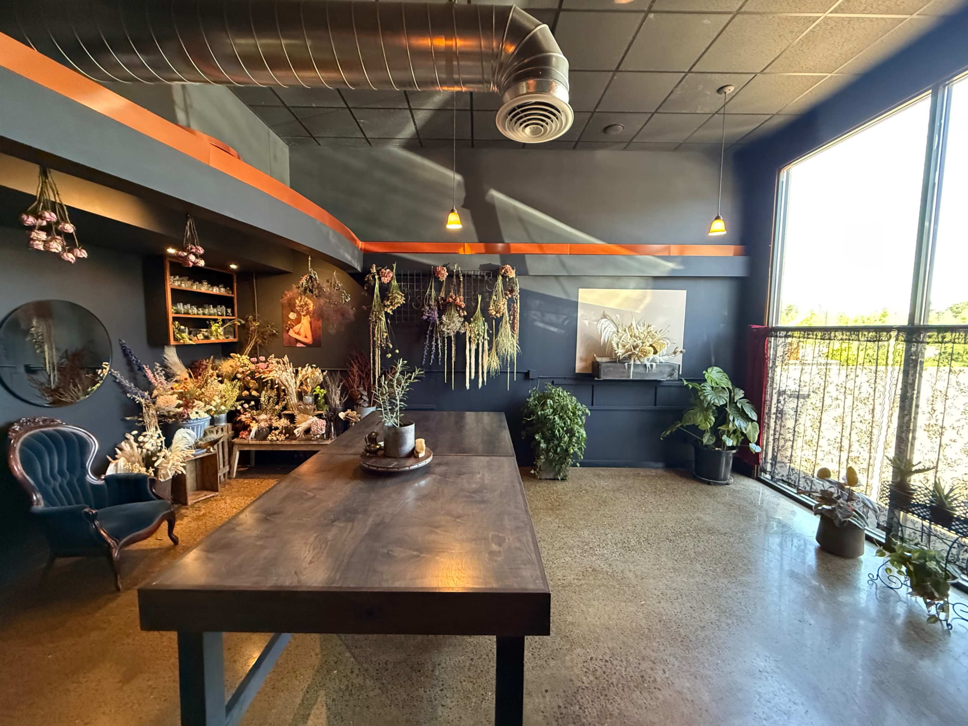The image shows a spacious interior of a room featuring a large wooden table, decorative plants, and various floral arrangements on the walls and shelves.