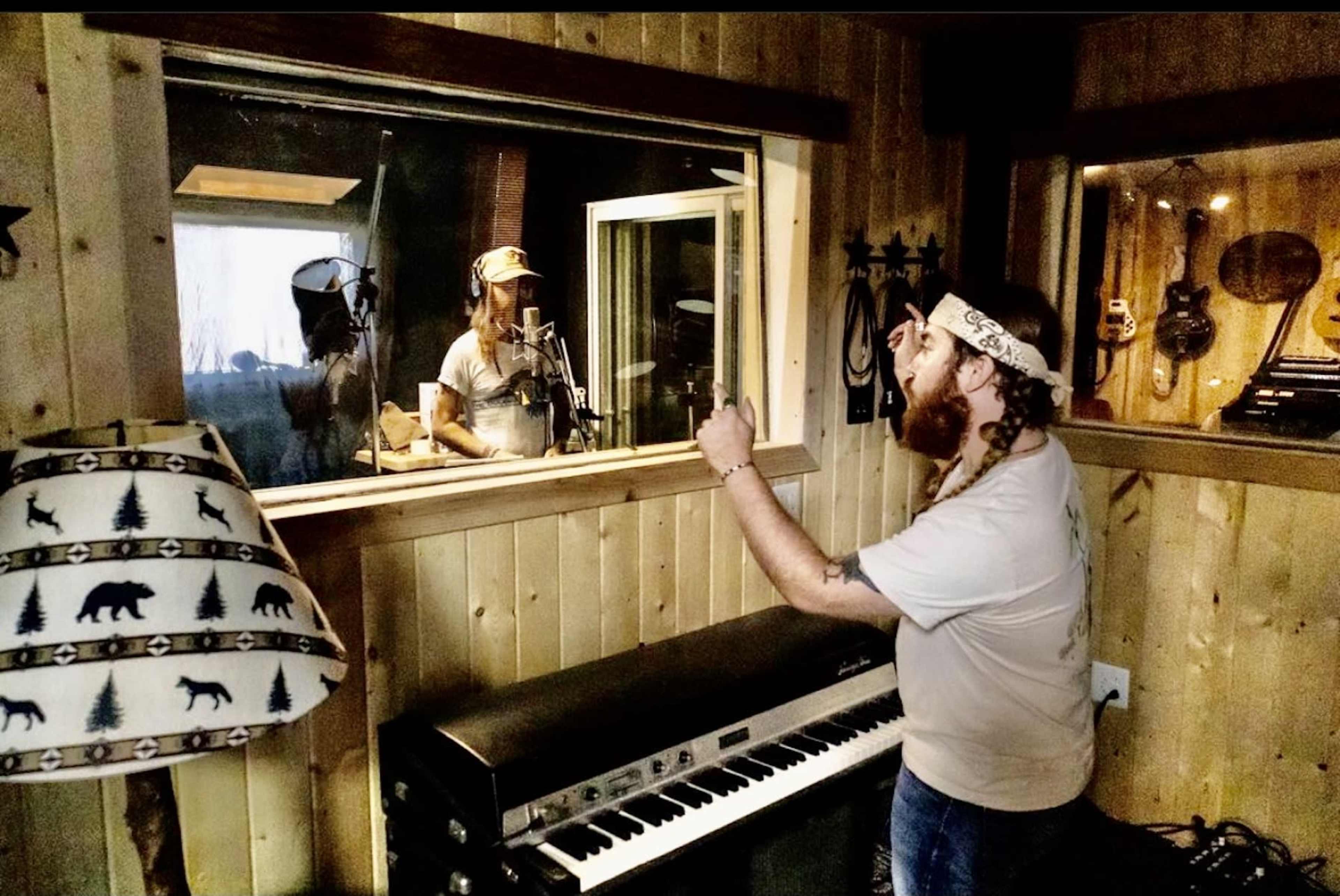 A musician stands in a recording studio, gesturing toward another artist behind a glass sound booth.