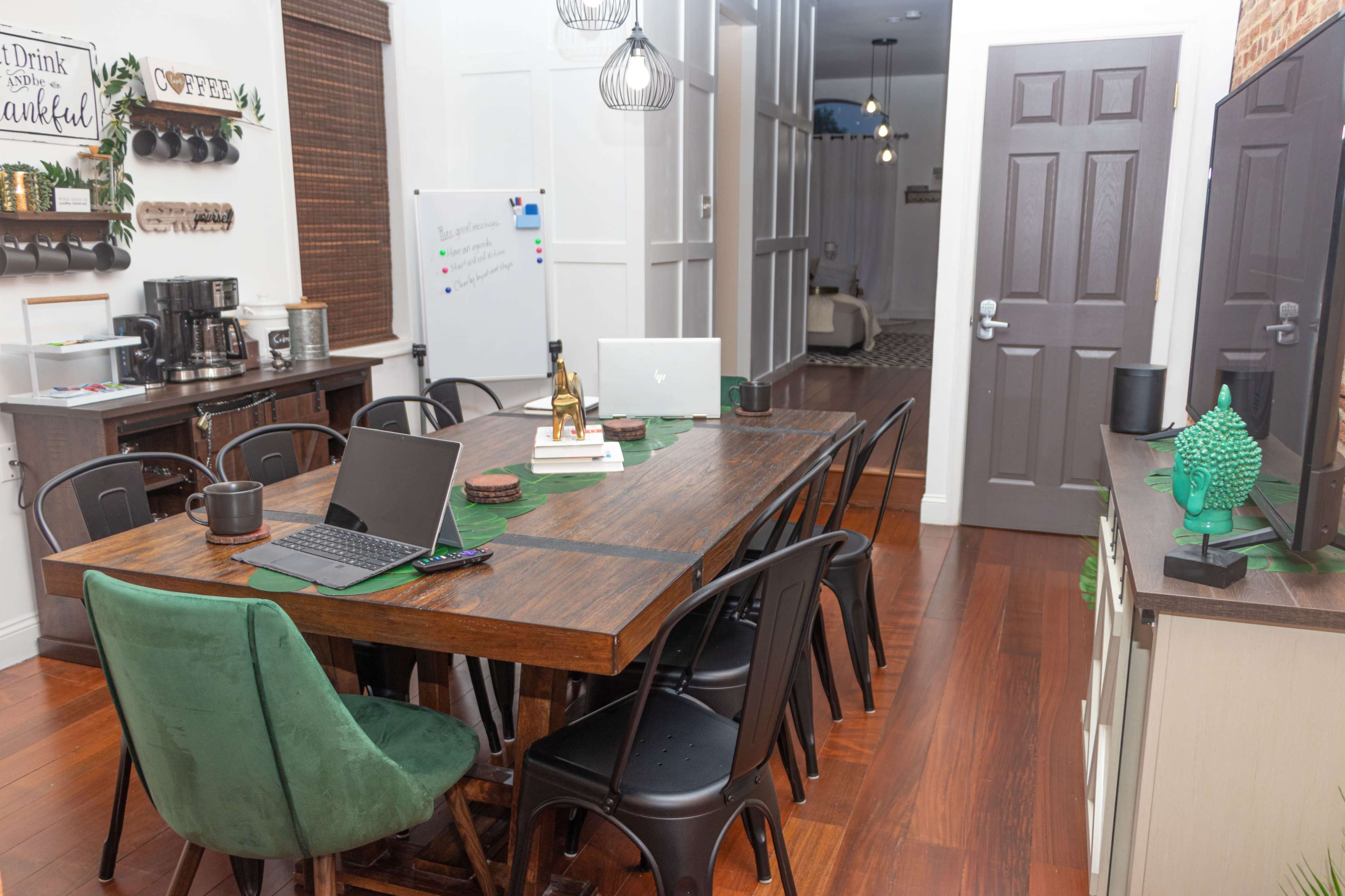 A wooden dining table with laptops and a coffee cup is set in a well-lit kitchen area featuring dark cabinetry and a doorway leading to another room.