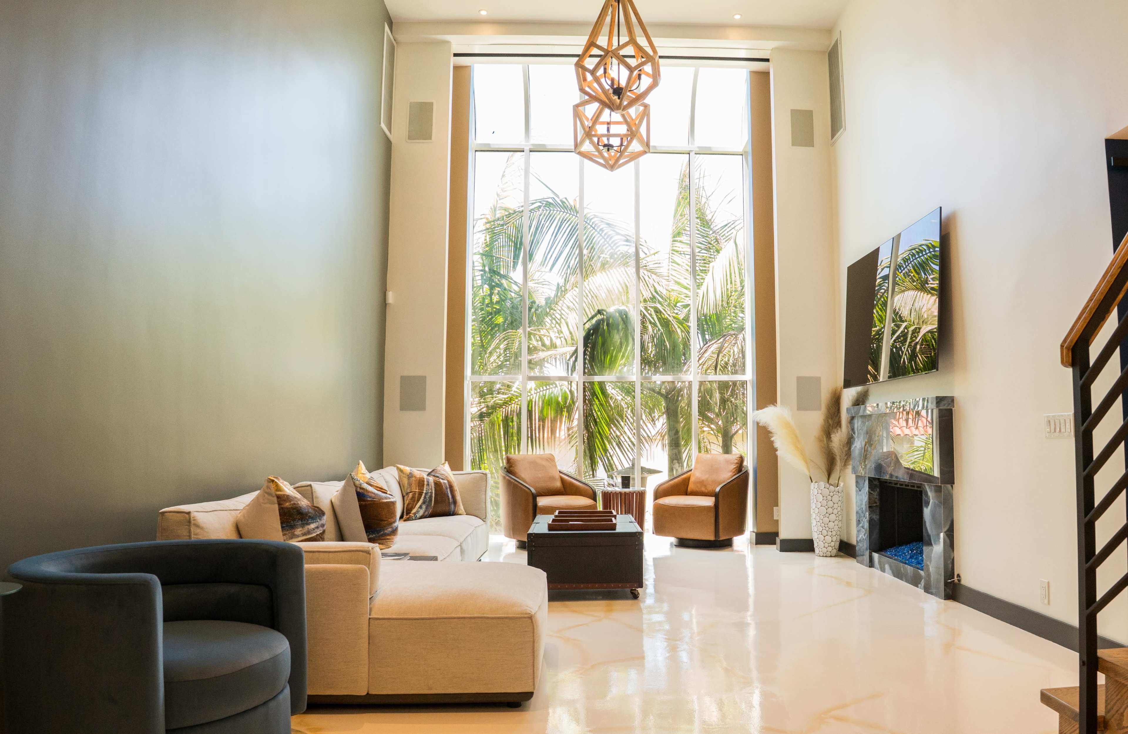 A modern living room features a large window with palm trees visible outside, a sectional sofa, two armchairs, and a coffee table, all arranged around a fireplace.