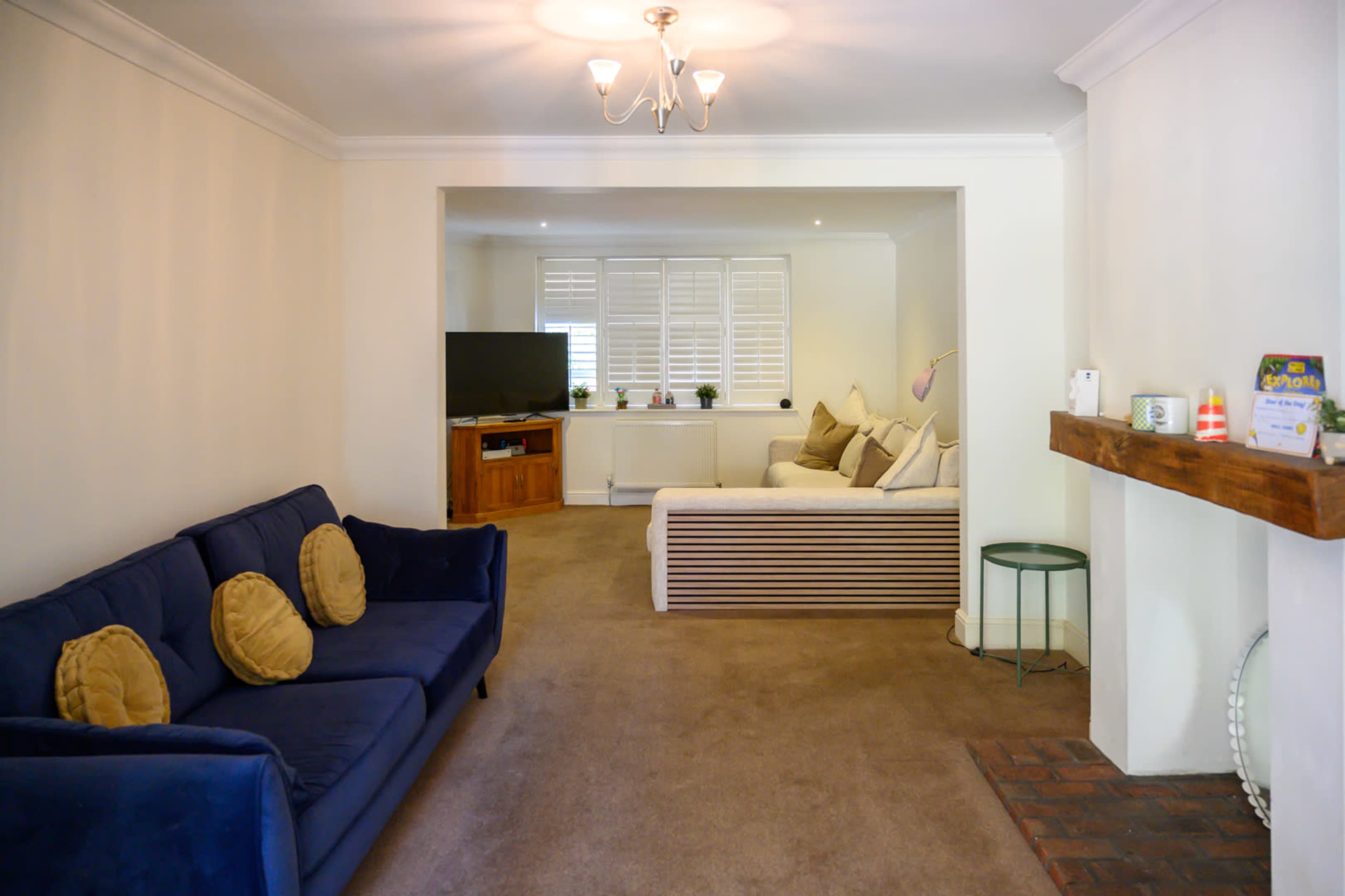 The image shows a living room with a blue sofa, a wooden coffee table, a TV mounted on the wall, and a light-filled area with a white couch and decorative items on a mantel.