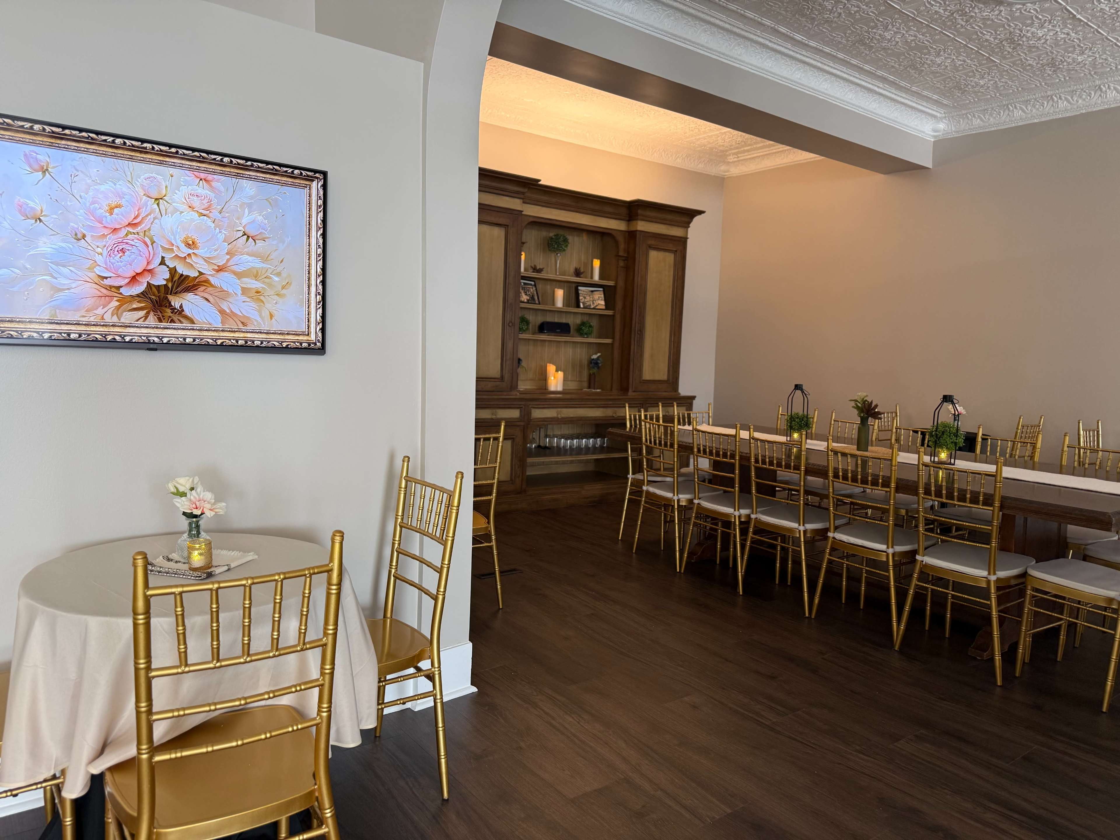 The image shows a decorated dining area with golden chairs and tables set up for an event, featuring a floral painting on the wall.