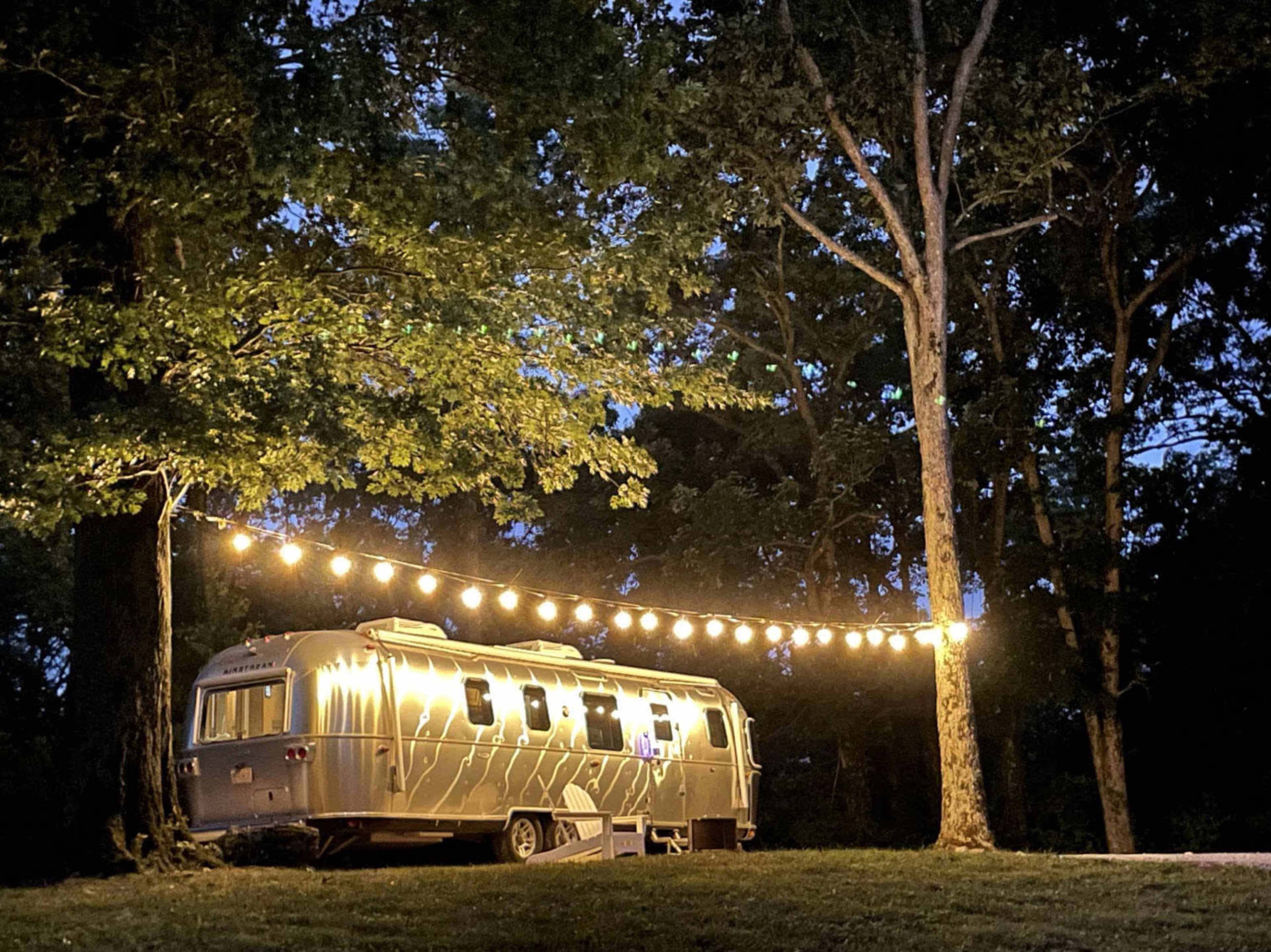 An Airstream trailer is parked under trees illuminated by string lights during the evening.