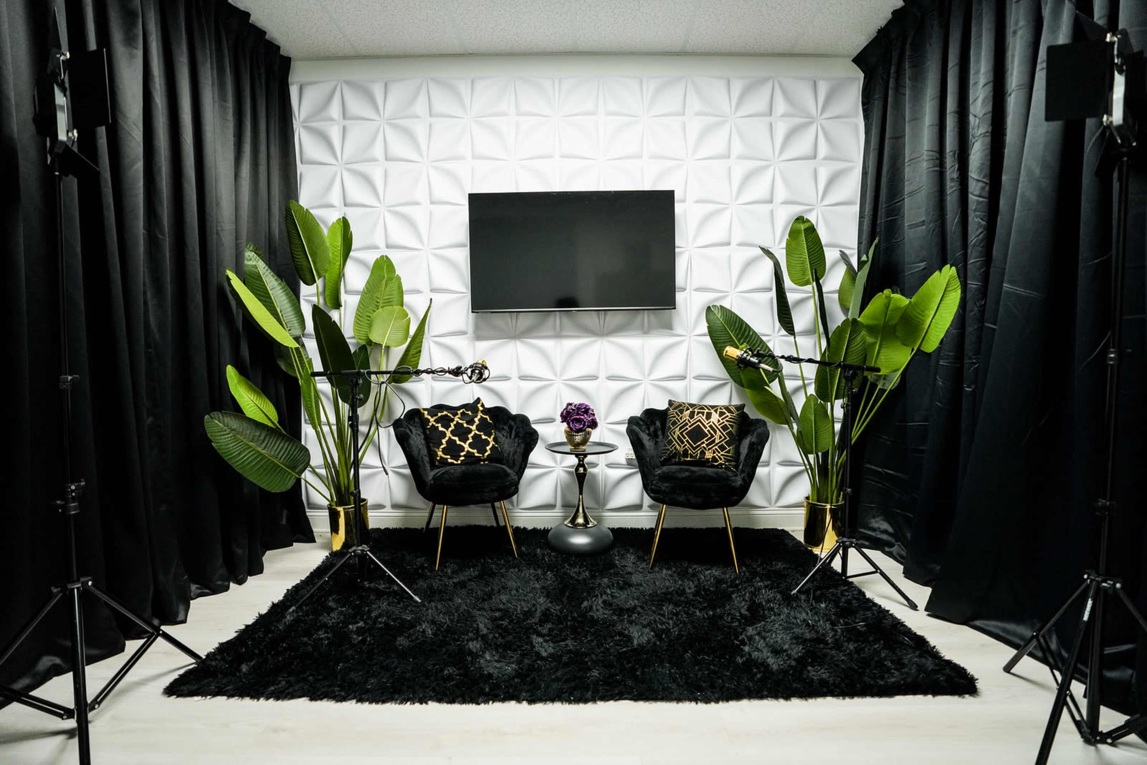 The image depicts a stylish interior setup with two black chairs, a small table, a large plant, and a wall-mounted television, all framed by black curtains and a textured white wall.
