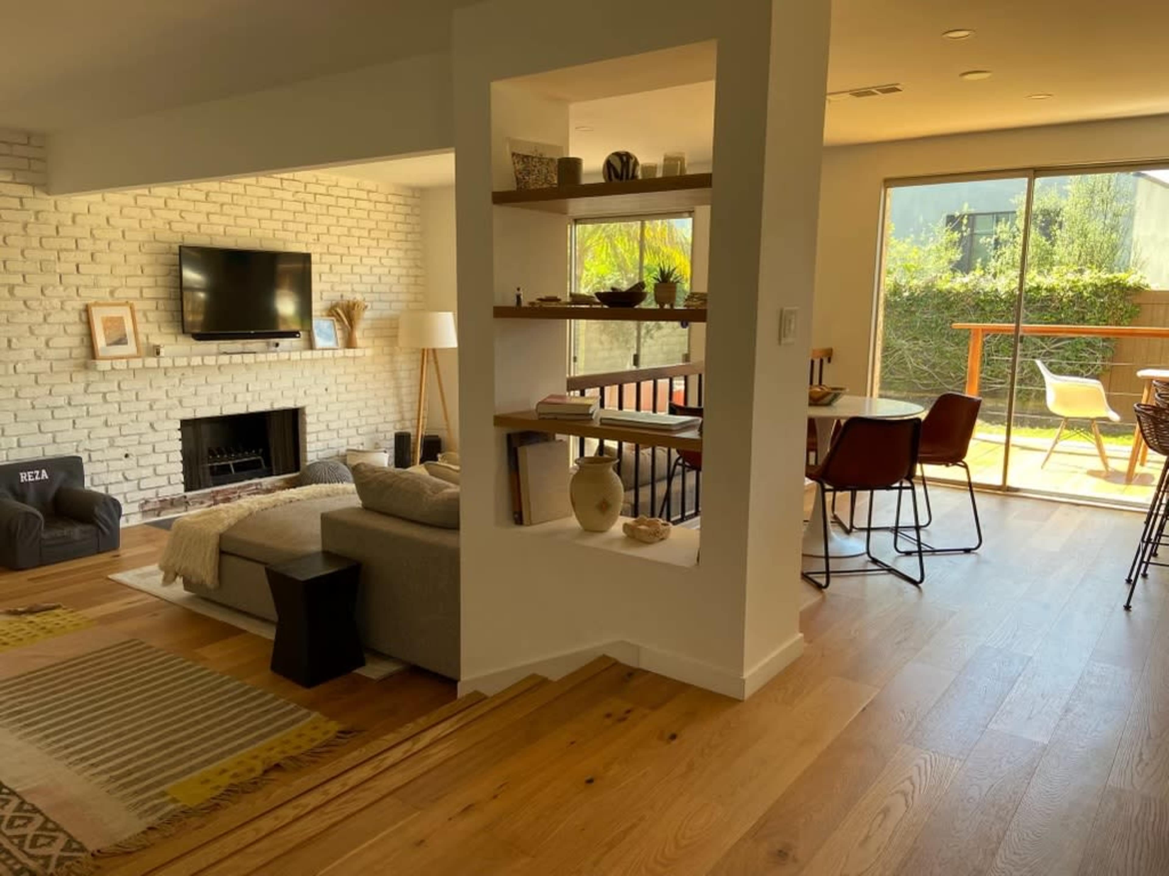 A minimalist living space with a gray sectional sofa, a brick accent wall, a small shelf with decorative items, and a large window leading to an outdoor patio.