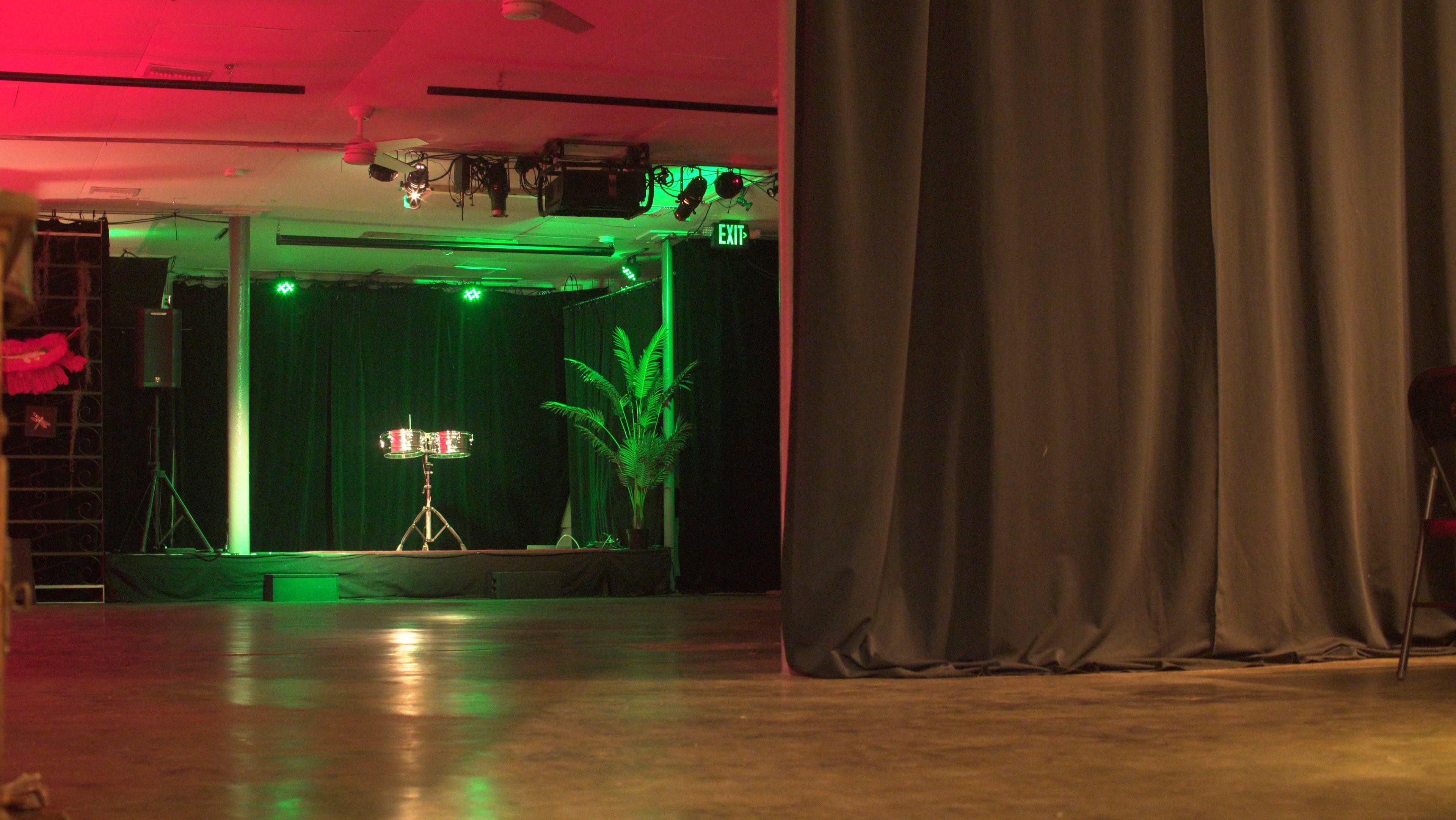 The image shows a dimly lit performance space with green lighting, featuring a small drum set on a stage behind a black curtain.