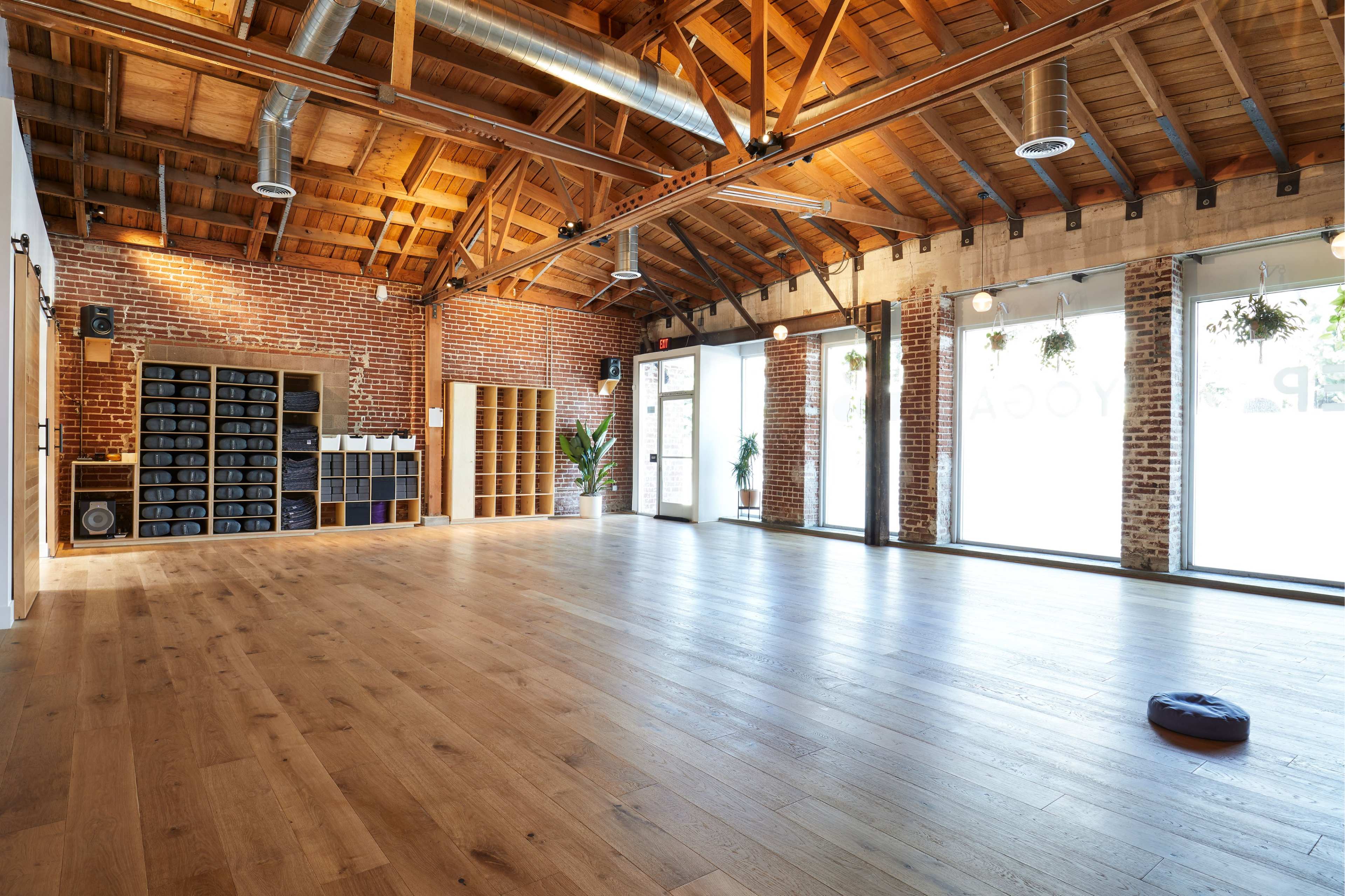 A spacious yoga studio with wooden floors, high ceilings, exposed brick walls, and large windows allowing natural light to enter.