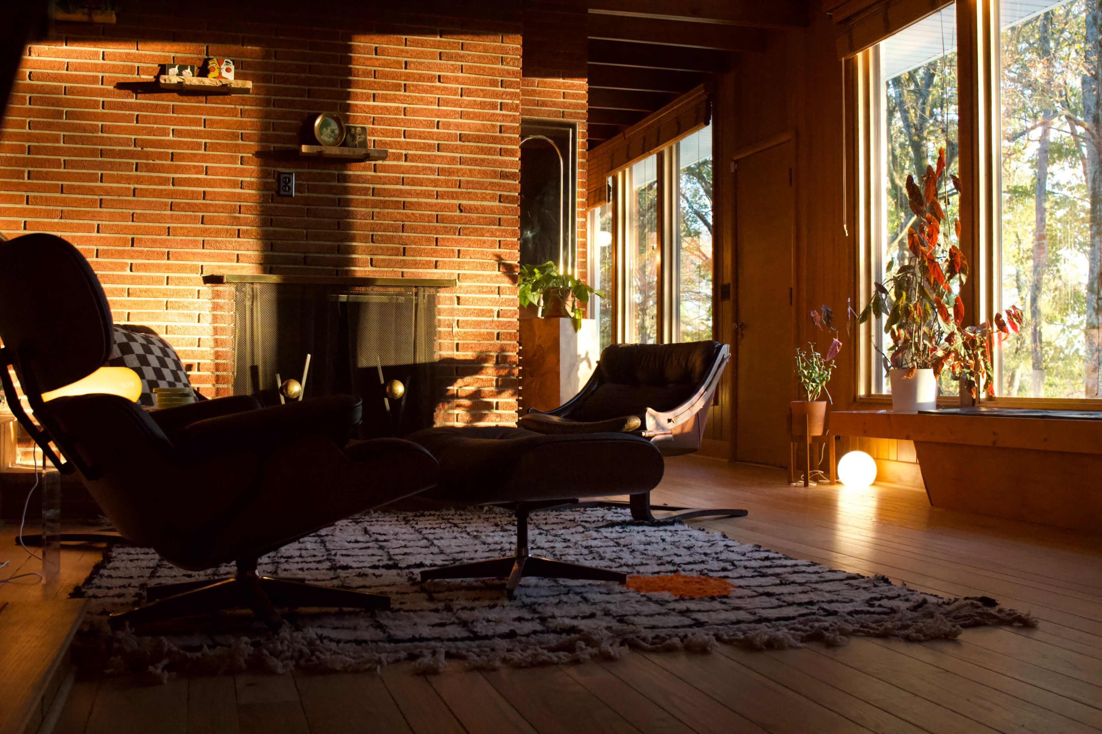 A cozy living room features a brick wall, large windows letting in natural light, and mid-century modern furniture arranged around a textured rug.