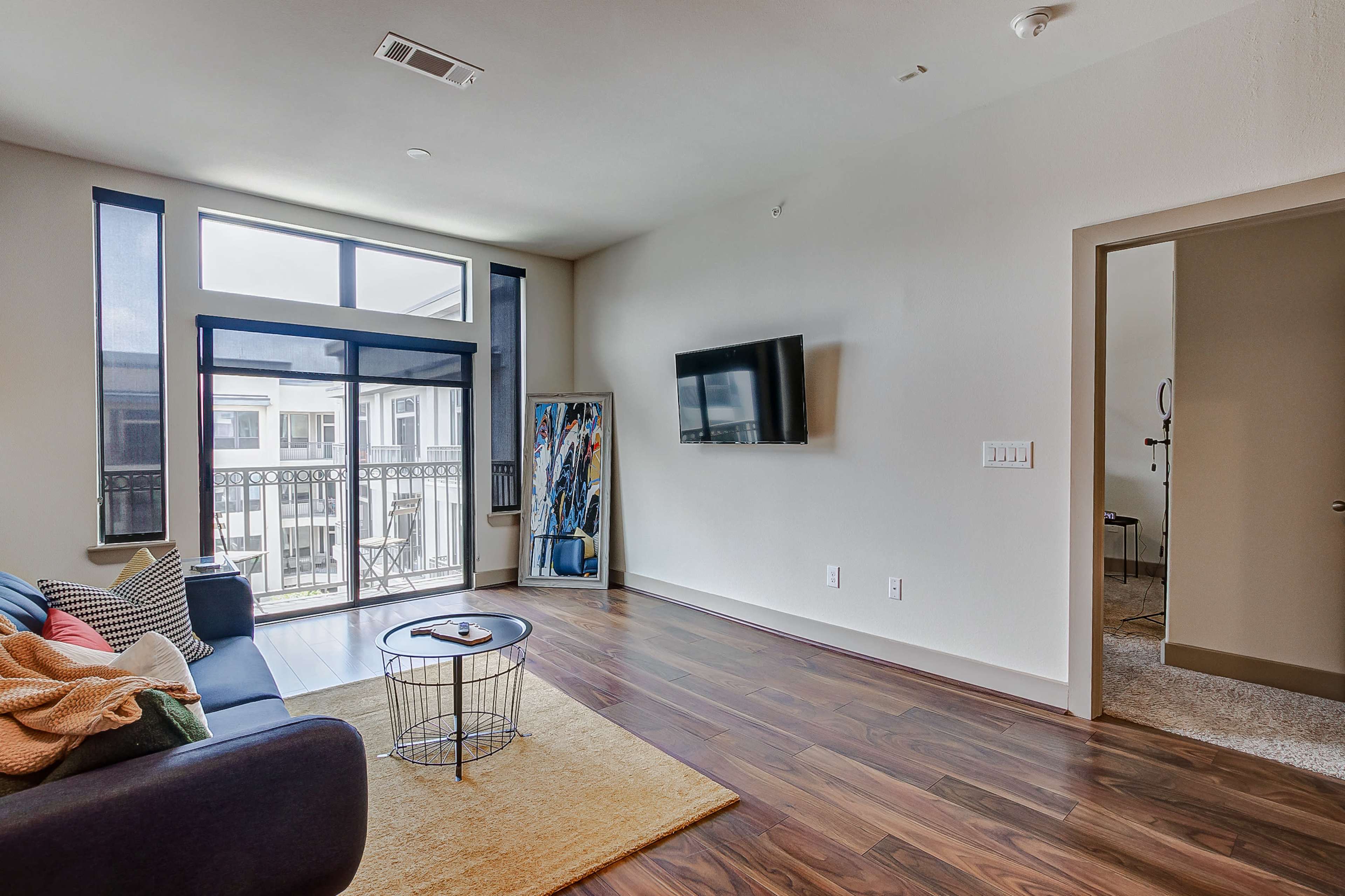 A living room features a blue sofa, a round coffee table, a wall-mounted television, large windows, and a doorway leading to another room.