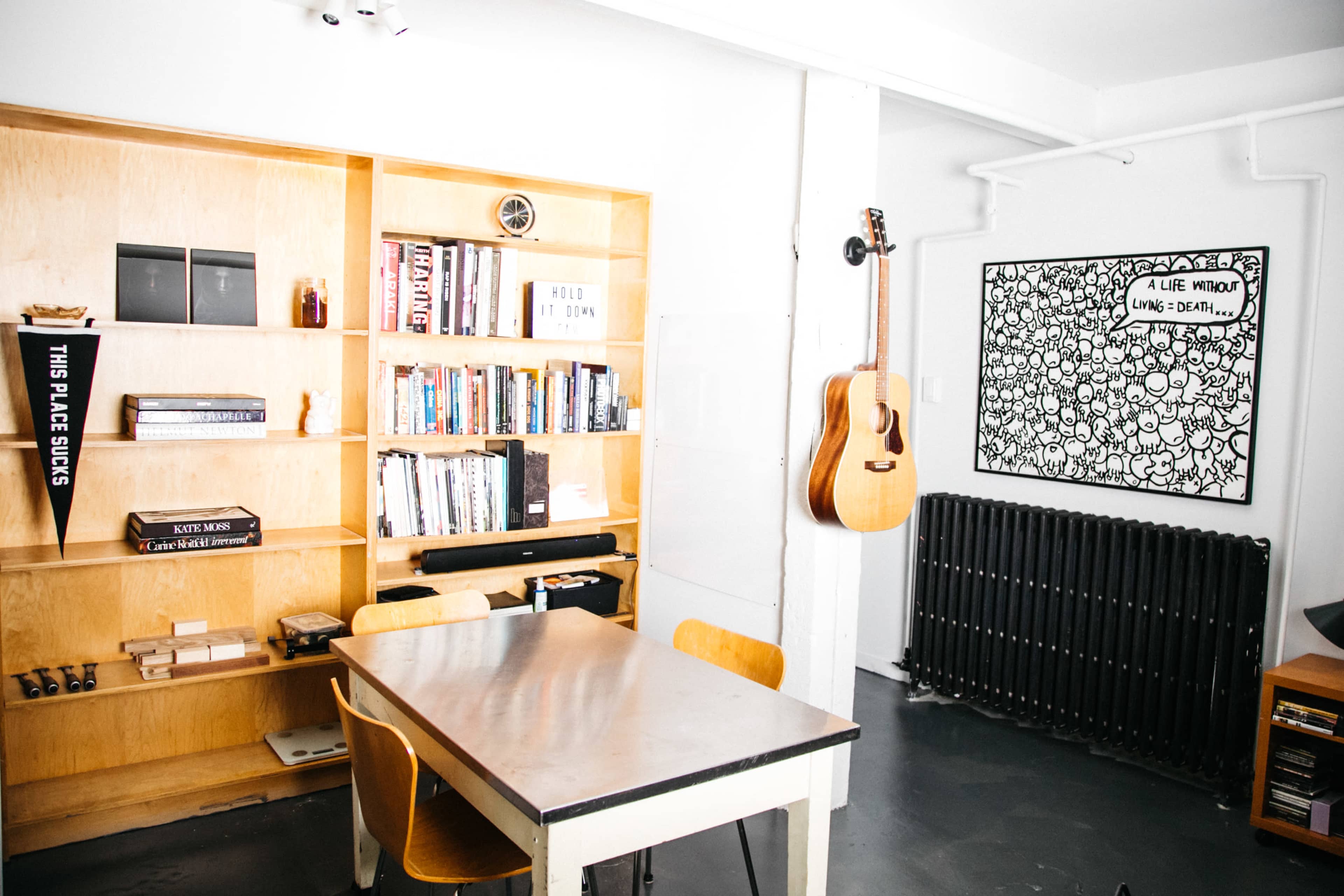The image shows a minimalist room with a wooden bookshelf filled with books, a table with four chairs, and a wall featuring an art piece and a guitar hanging next to a radiator.
