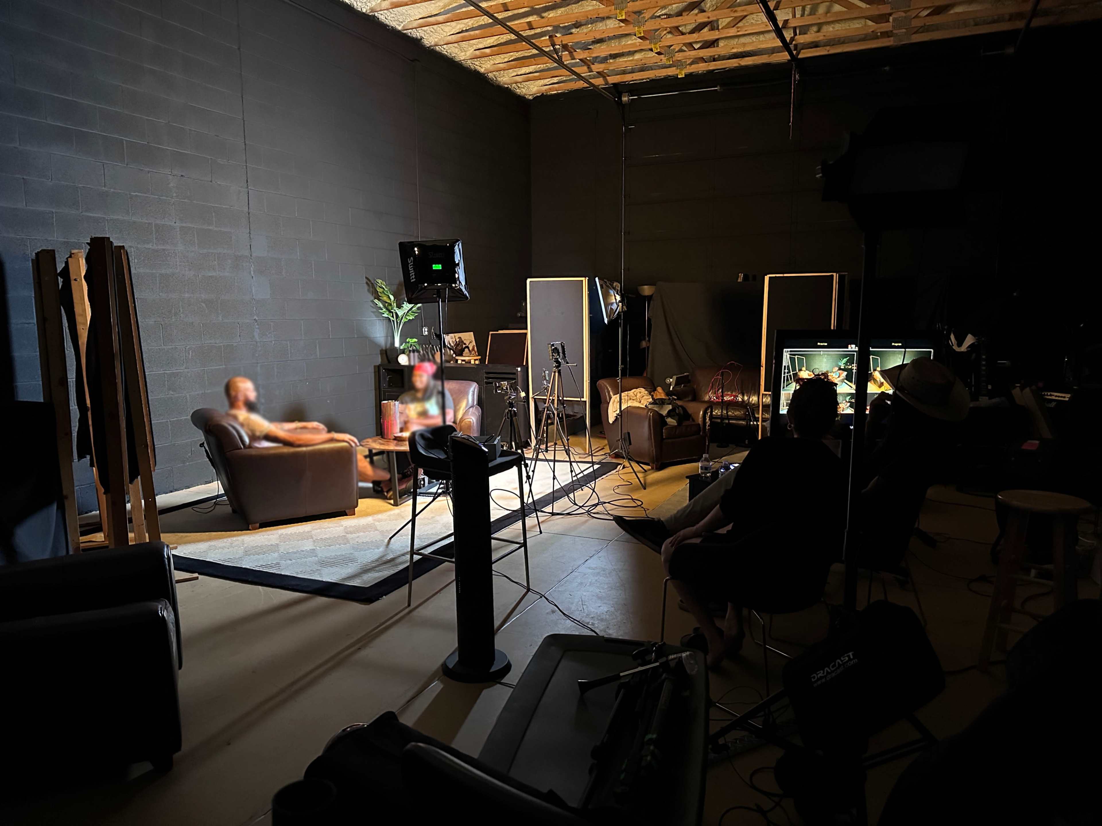The image shows a dimly lit studio with two people sitting on a couch, surrounded by cameras and lighting equipment.