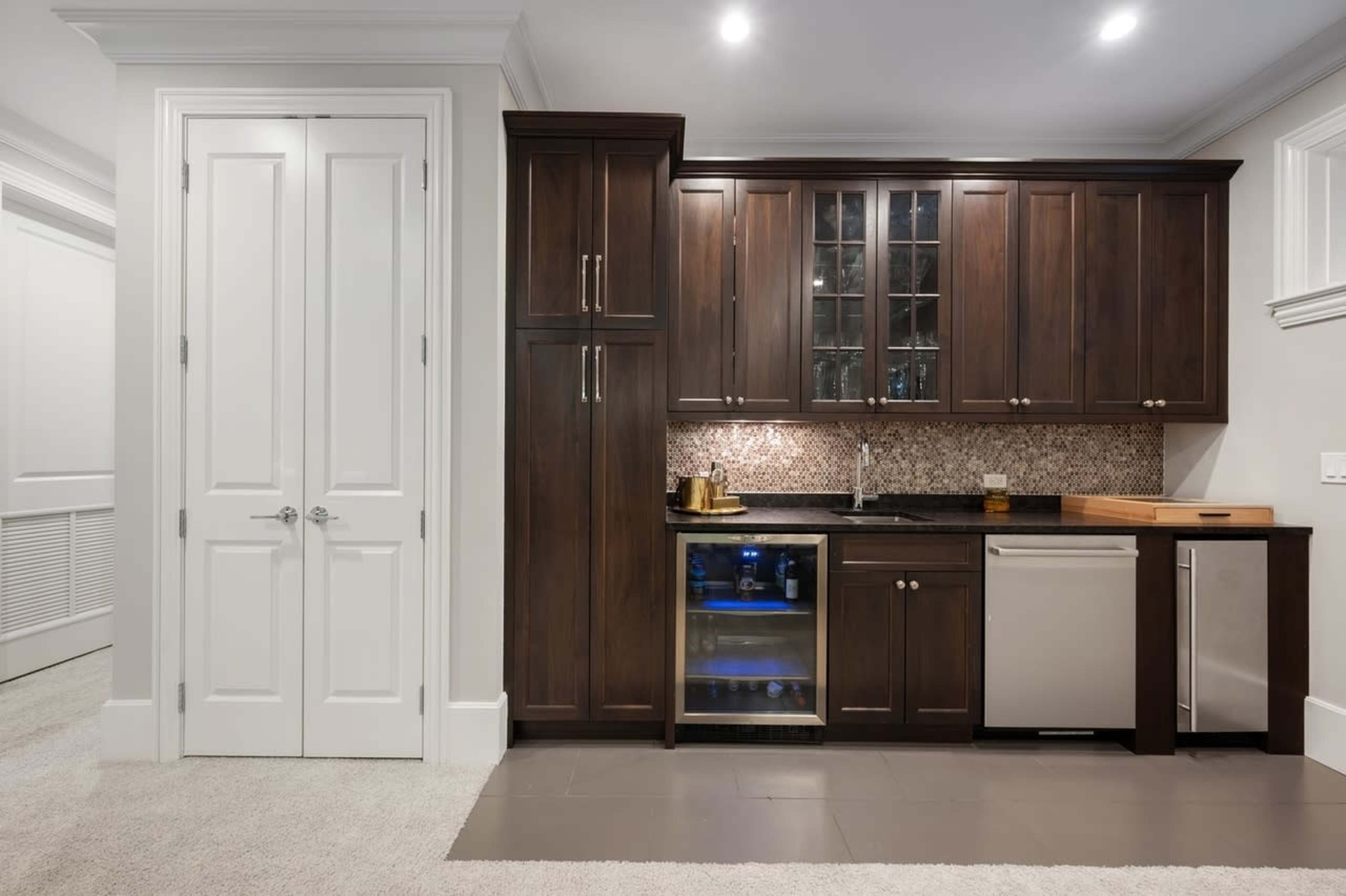 A modern kitchen area featuring dark wooden cabinetry, a small refrigerator, and a wine cooler beneath a countertop with a tile backsplash.