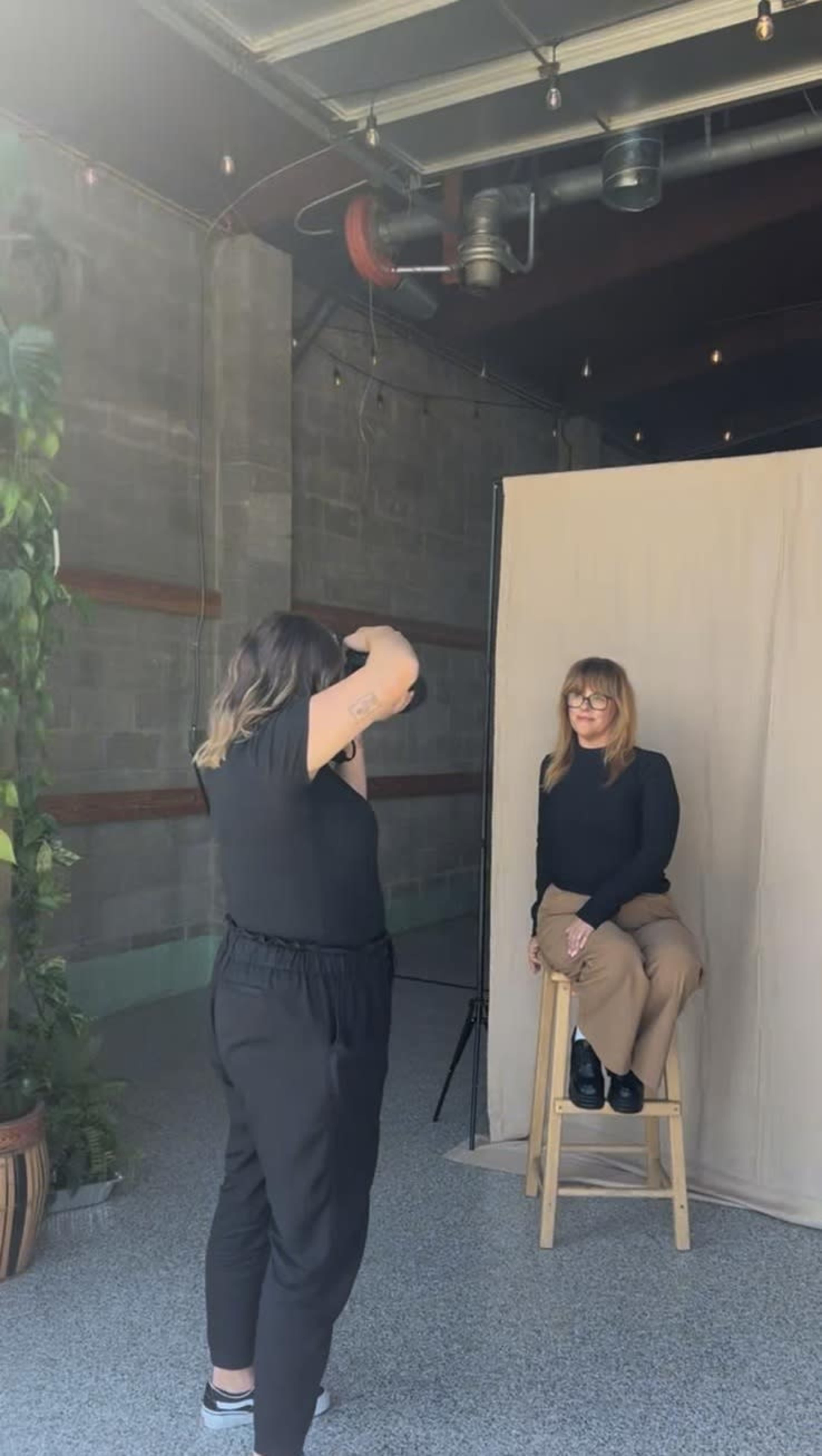 A photographer is taking a portrait of a seated woman against a neutral backdrop in a well-lit indoor space.