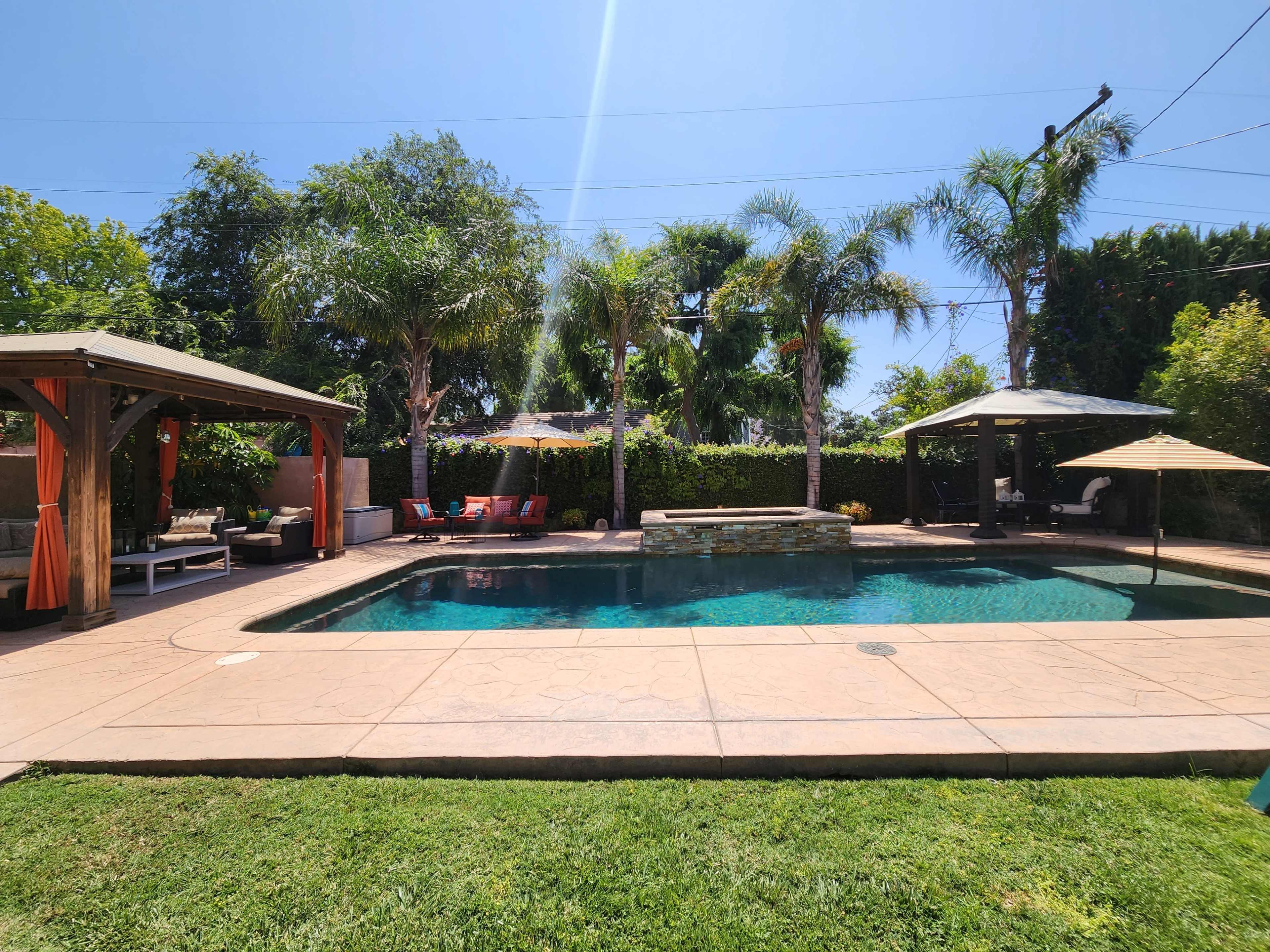 A backyard pool area with palm trees, two shaded seating structures, and lounge chairs beside a clear blue pool.