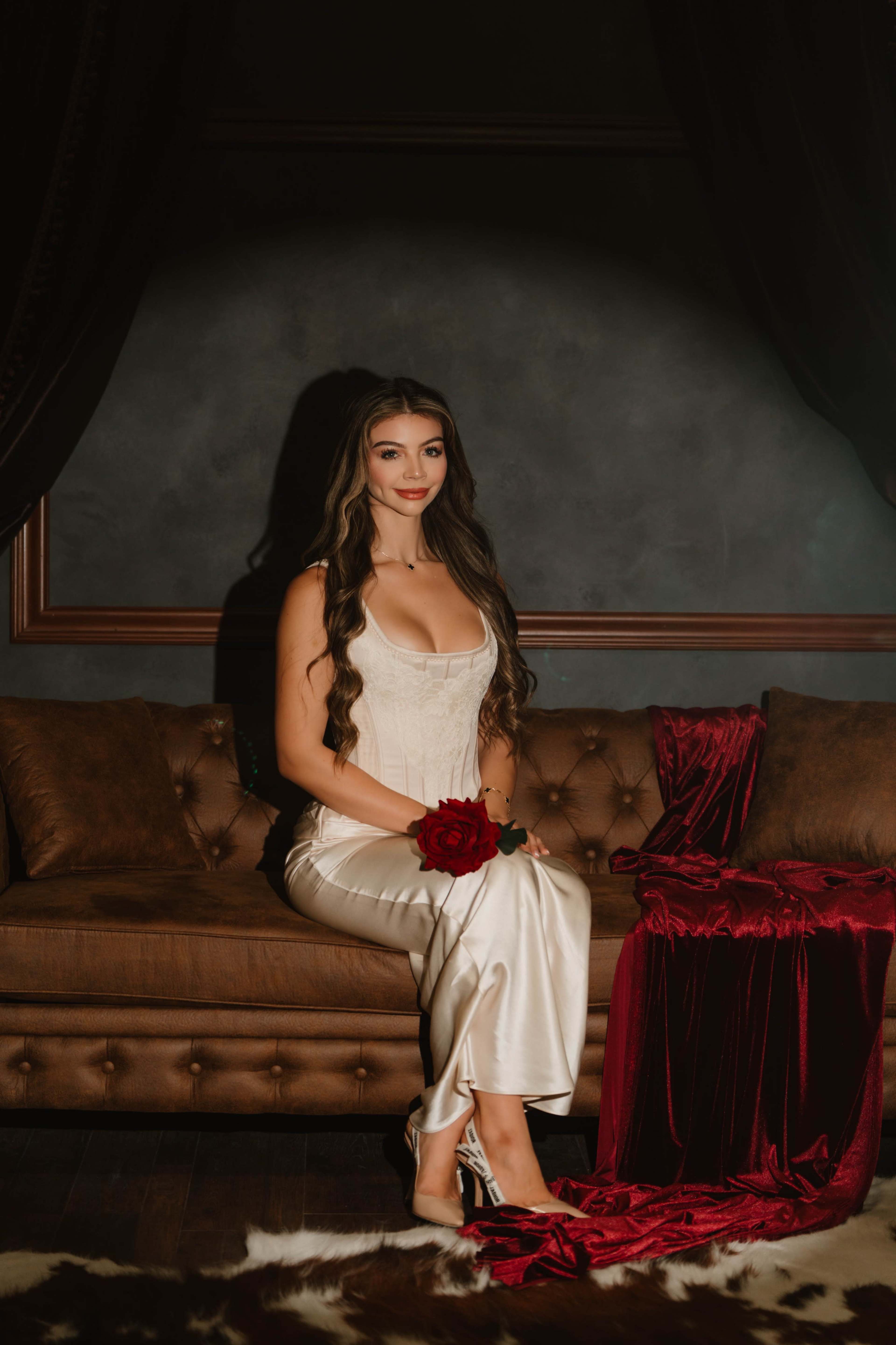 A woman in a satin dress sits on a brown couch, holding a red rose, with dark curtains and a plush red fabric in the background.