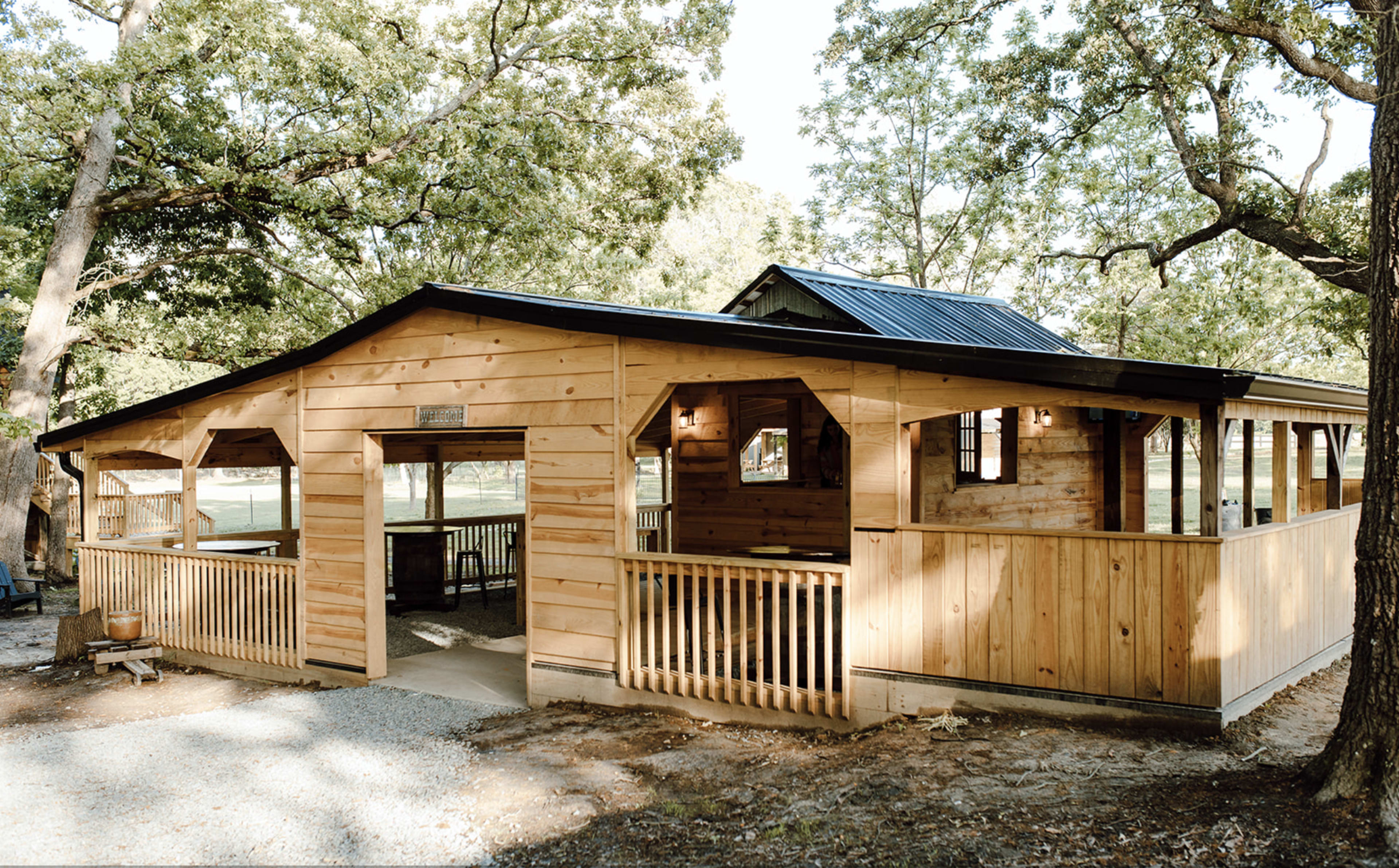 The image shows a large wooden structure with a sloped roof, featuring multiple openings and a porch area, surrounded by trees in a natural setting.