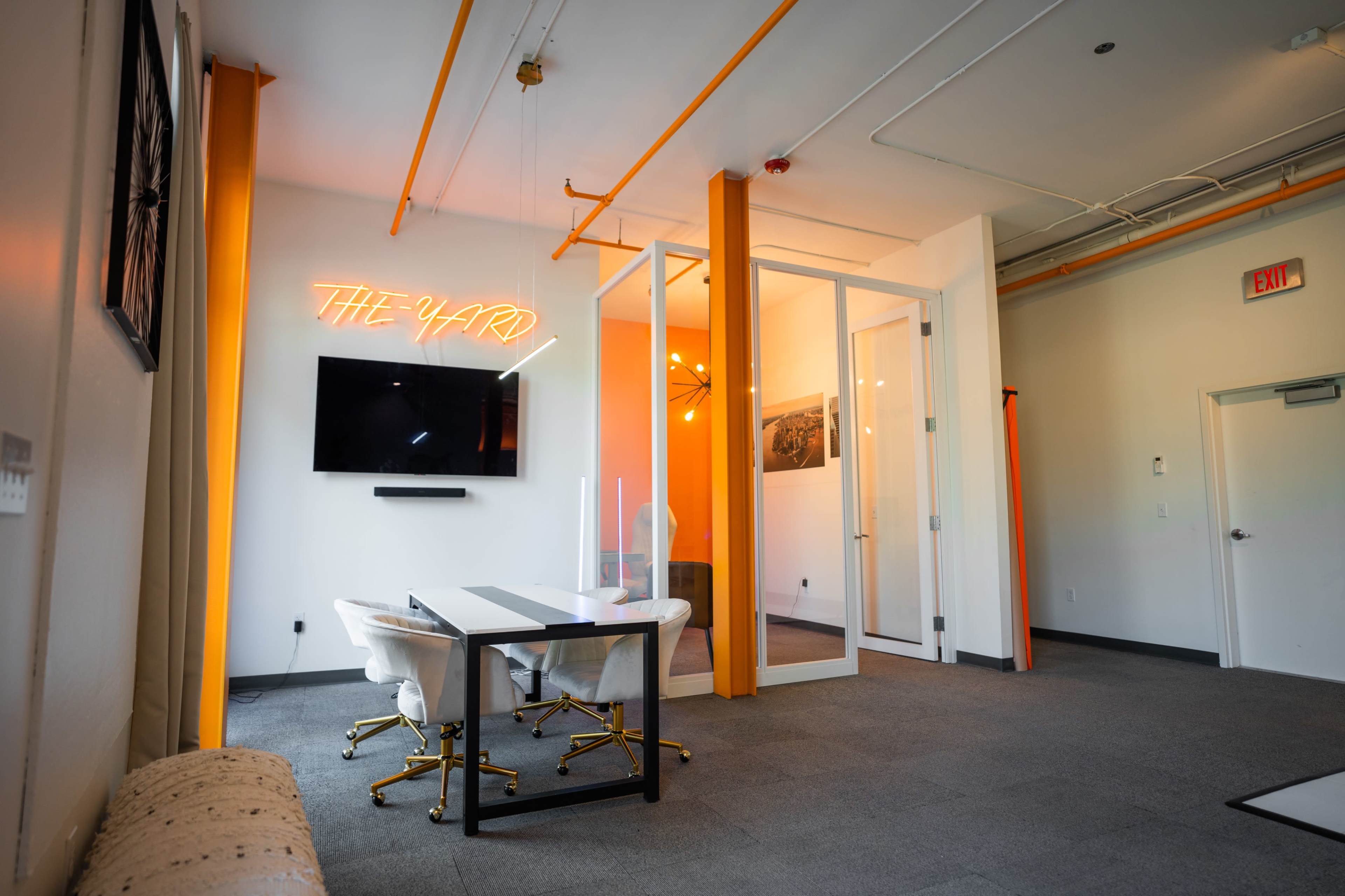 A modern office space features a meeting area with a glass-walled room, a wall-mounted TV, and bright orange accents.
