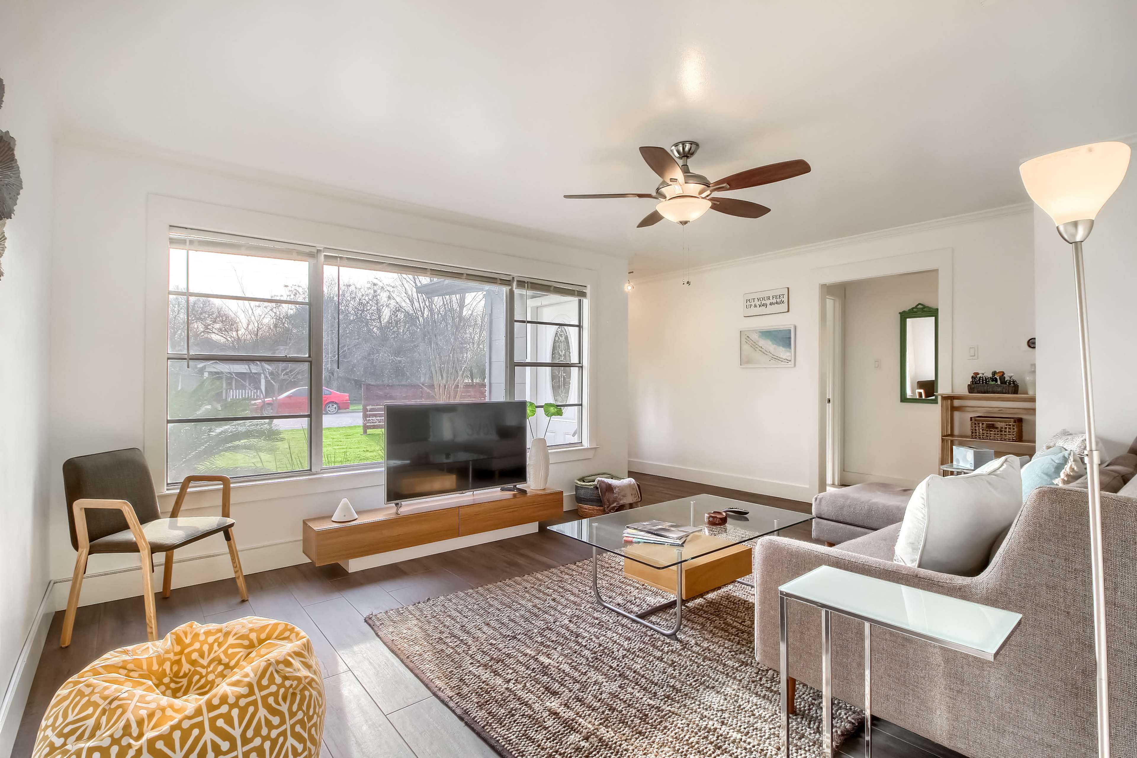 A spacious, well-lit living room features a large window, a comfortable sofa, a coffee table, and a ceiling fan.