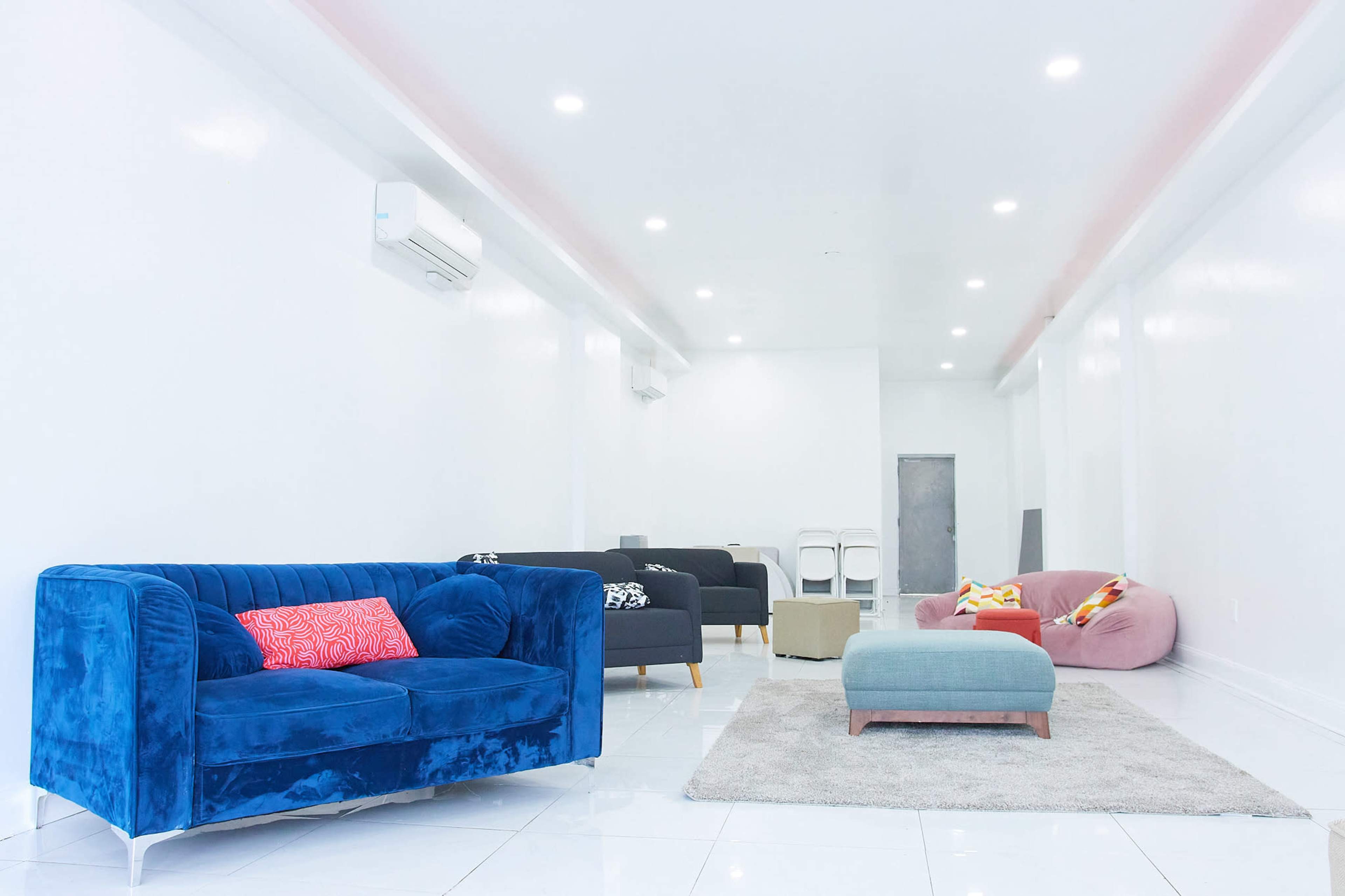 The image shows a modern, minimalist lounge area with a blue sofa, pink and gray seating, and white walls illuminated by bright ceiling lights.