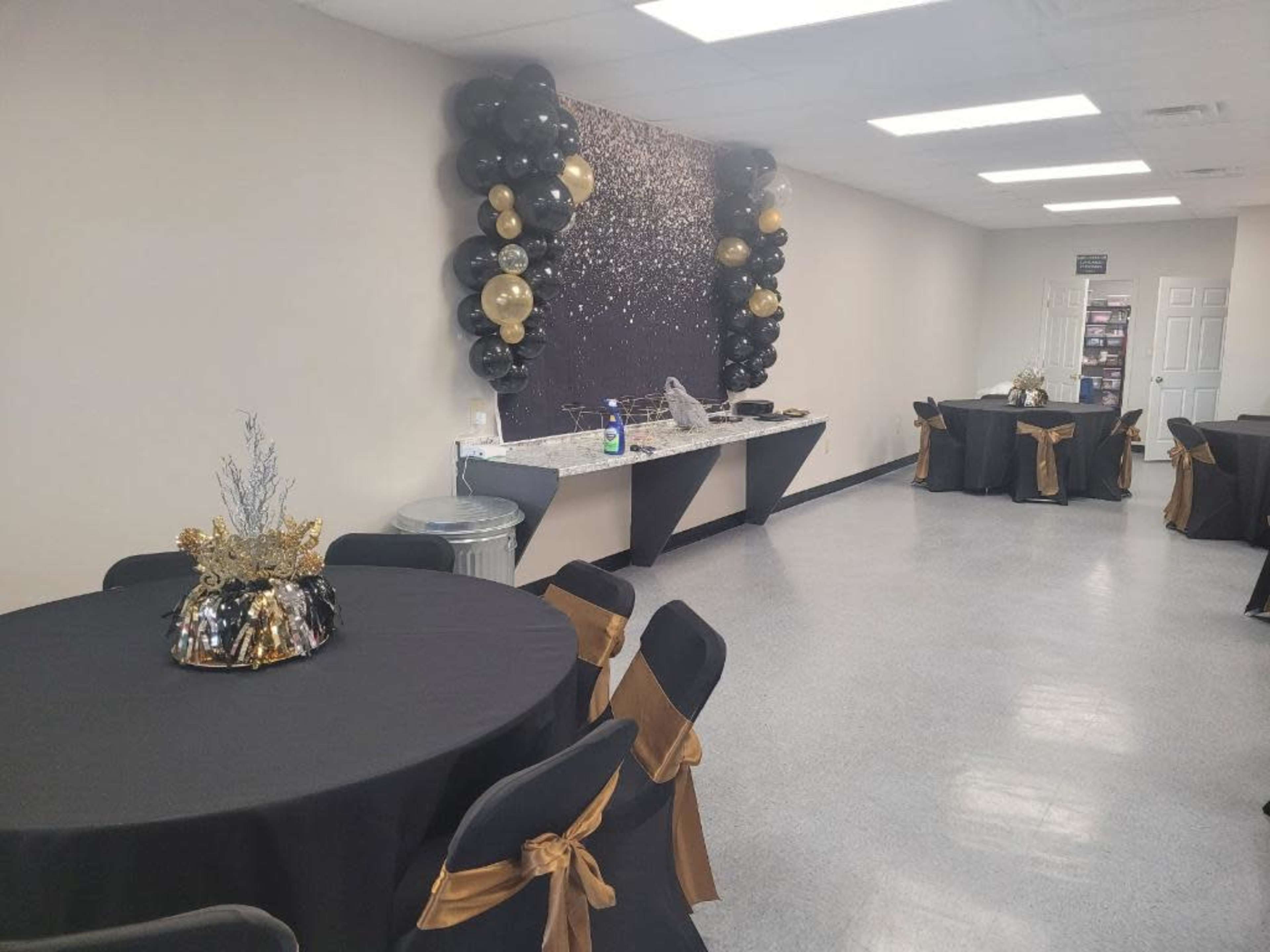 The image shows a decorated event space featuring black tablecloths with gold accents, a balloon backdrop, and a food preparation area along one wall.
