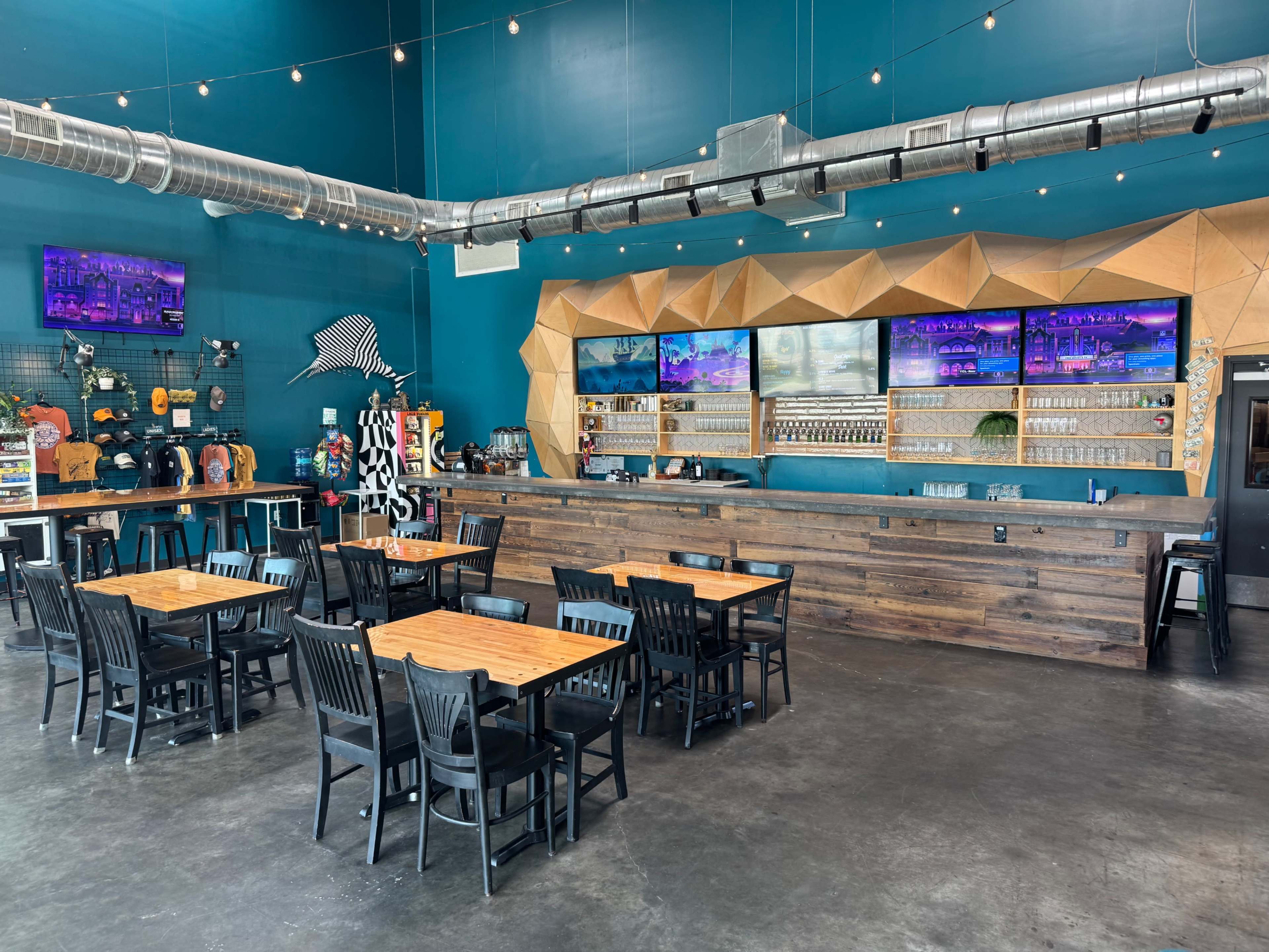 The image shows a spacious bar area with wooden tables and black chairs, featuring a large counter with several screens and a decorative wall behind it.