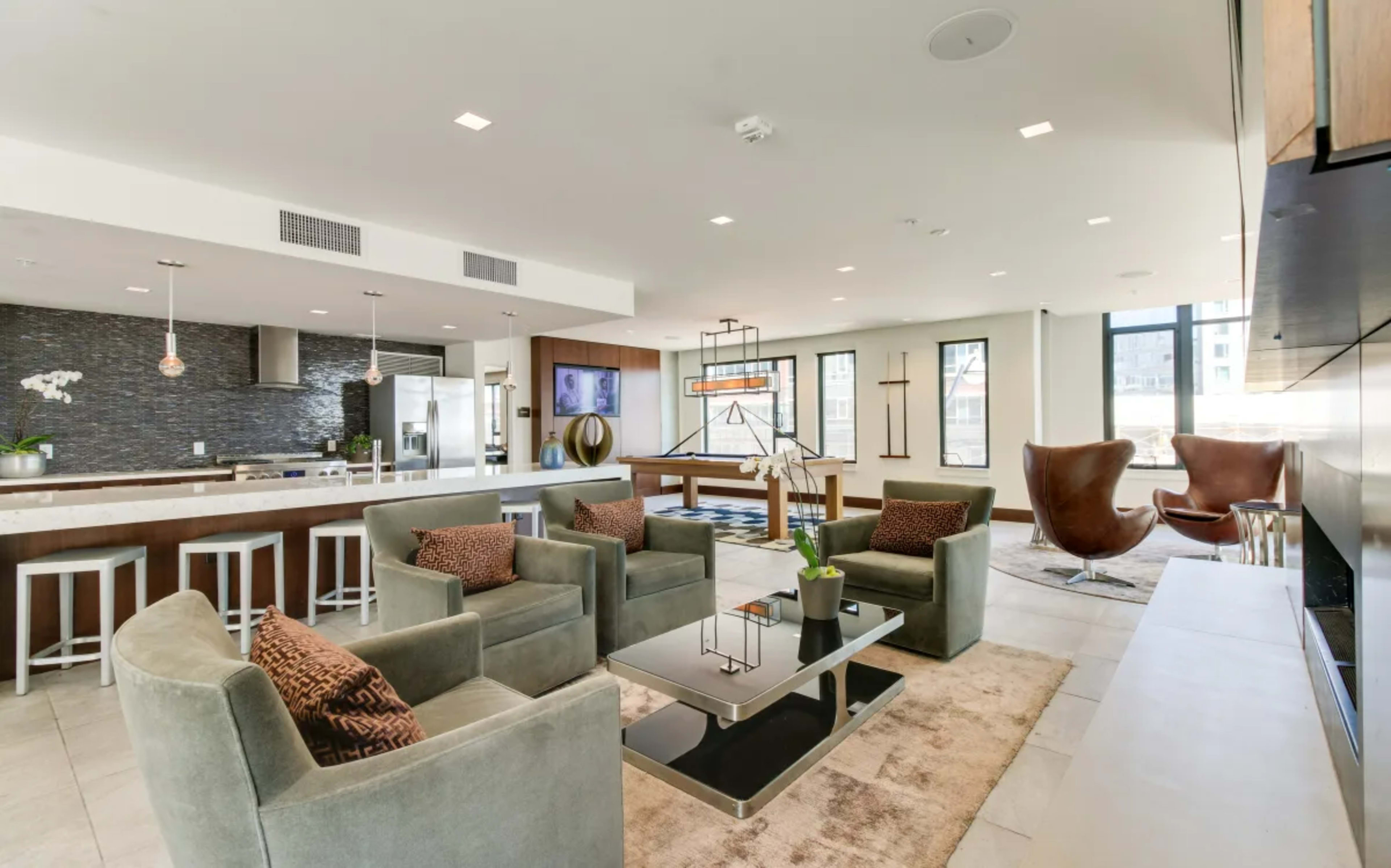 A modern living space features a kitchen area with bar seating, a large coffee table, comfortable seating arrangements, and large windows allowing natural light.