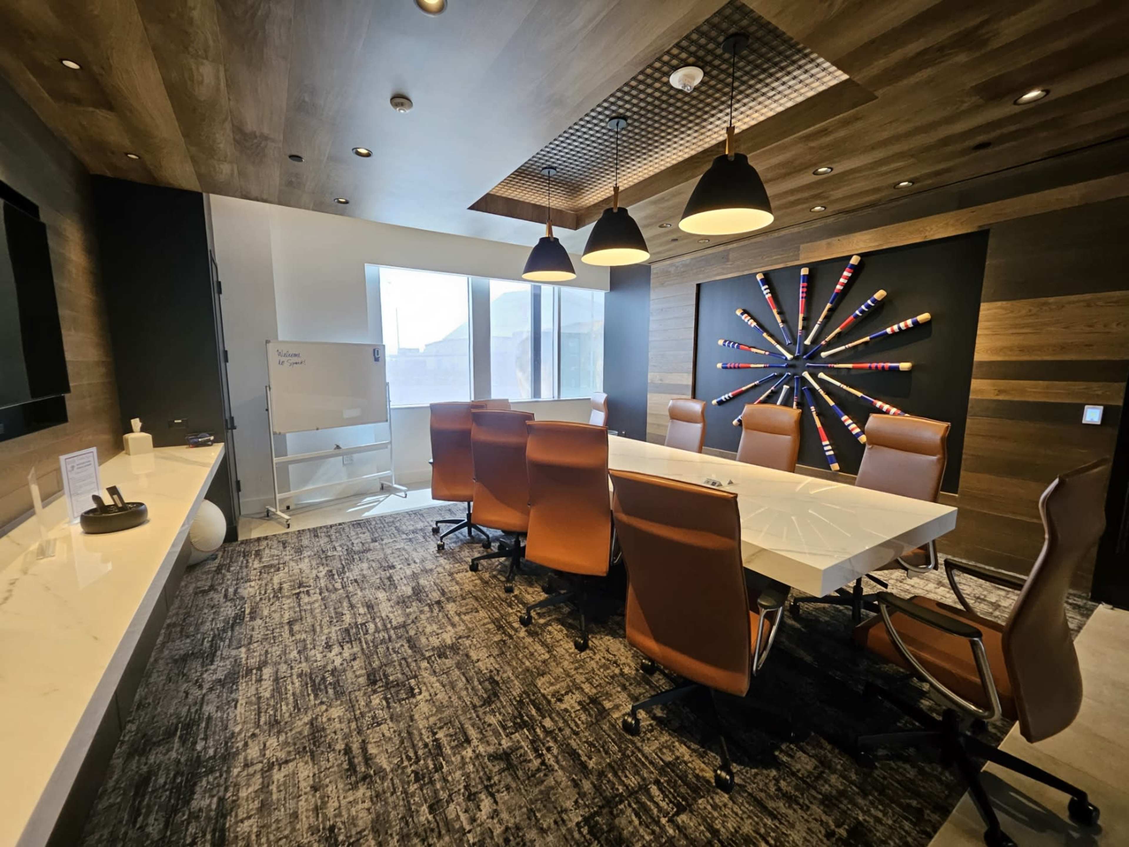 A modern meeting room features a large white conference table surrounded by brown leather chairs, with a colorful wall art installation and large windows providing natural light.