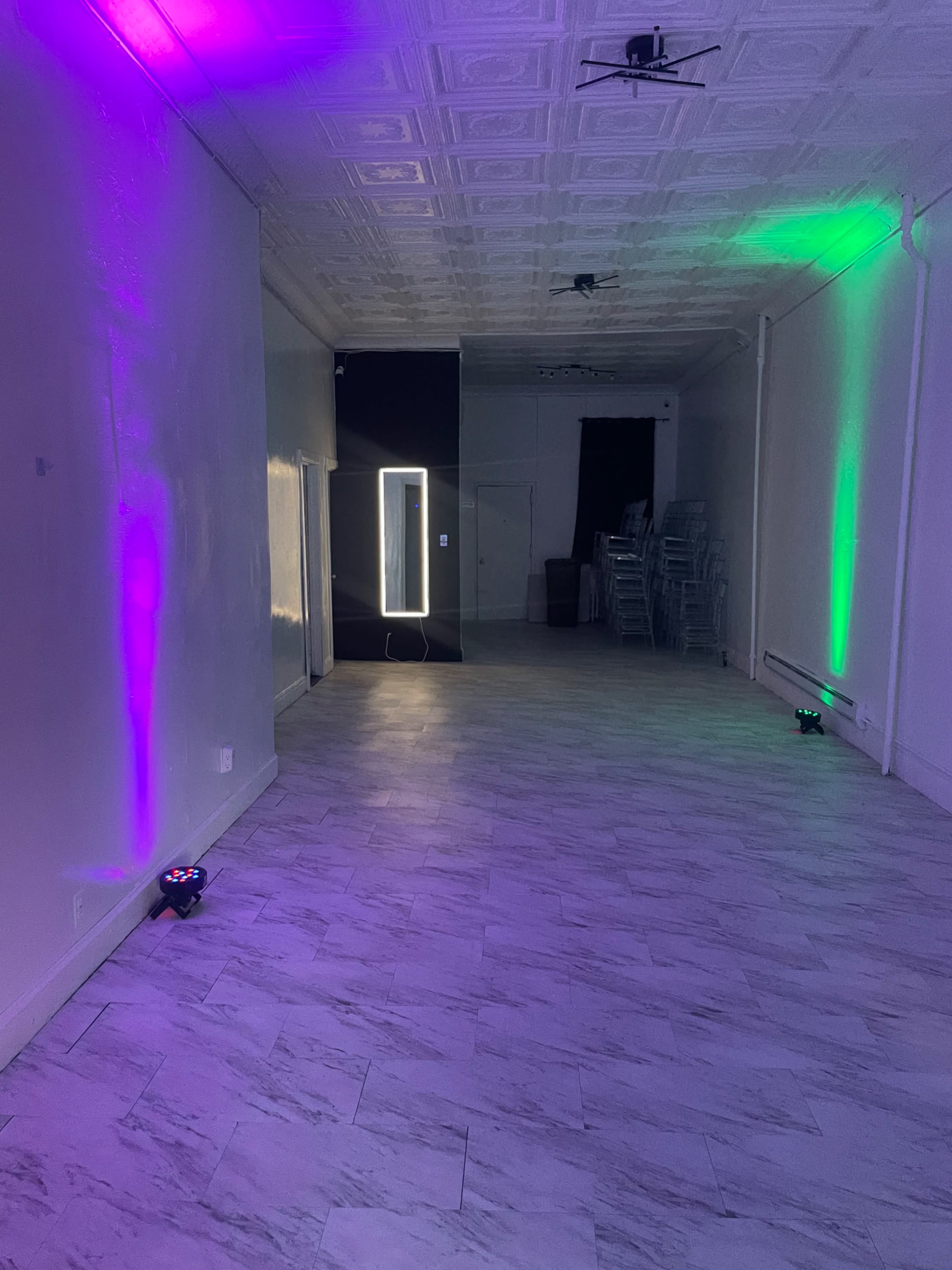 The image shows a spacious room with a tiled floor, featuring colored lights casting purple and green hues on the walls, and a mirror located near the back.