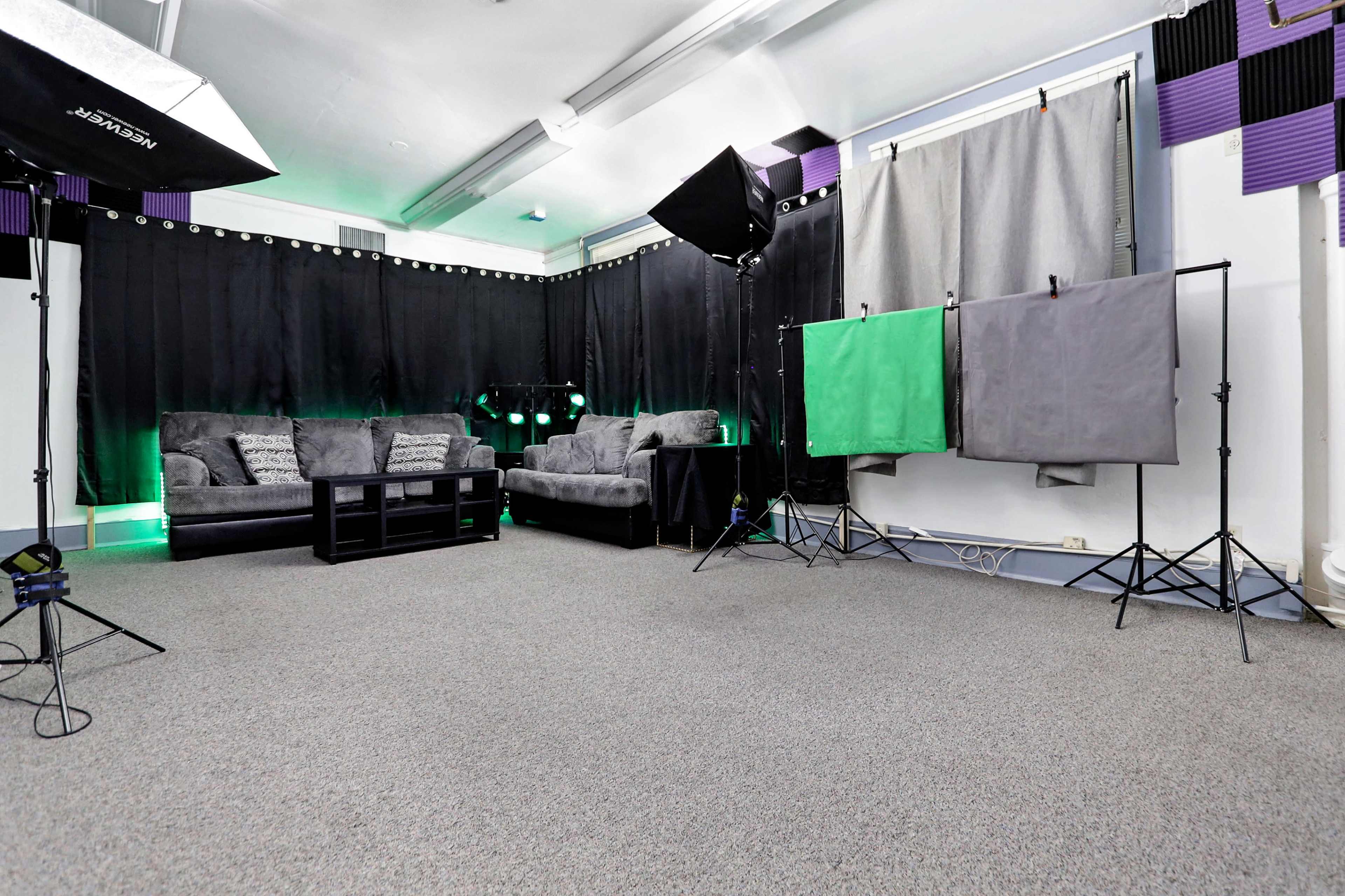 A studio setup with two gray and one green backdrop on stands, a sofa set, and lighting equipment in a carpeted room.