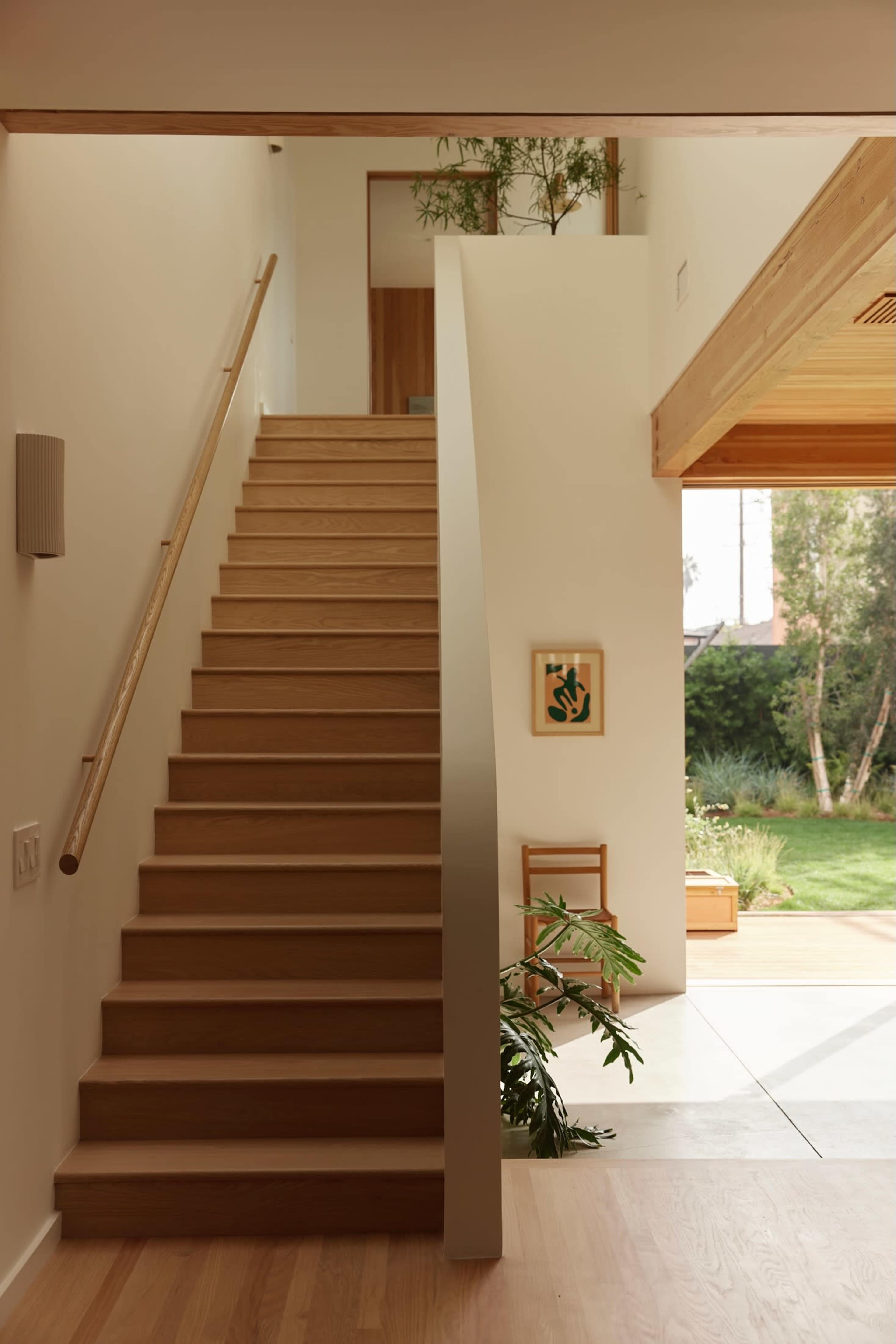 A wooden staircase leading to an upper level in a modern interior with large windows and greenery visible outside.