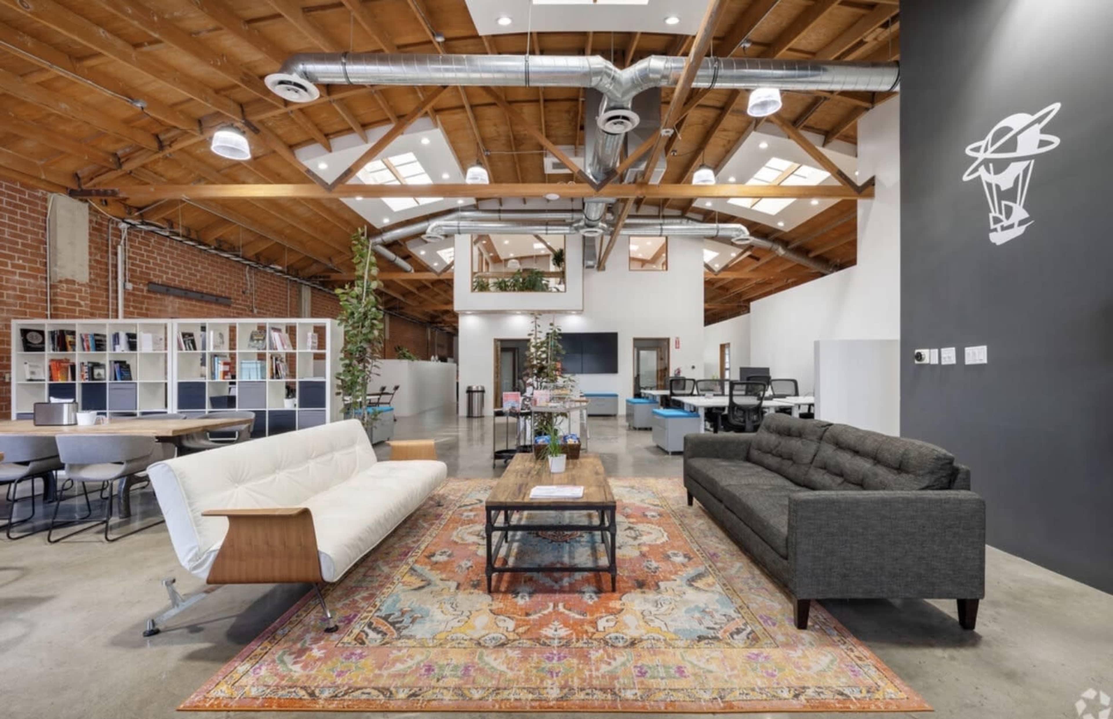 The image shows a modern office interior featuring two sofas, a decorative area rug, and exposed wooden beams on the ceiling.