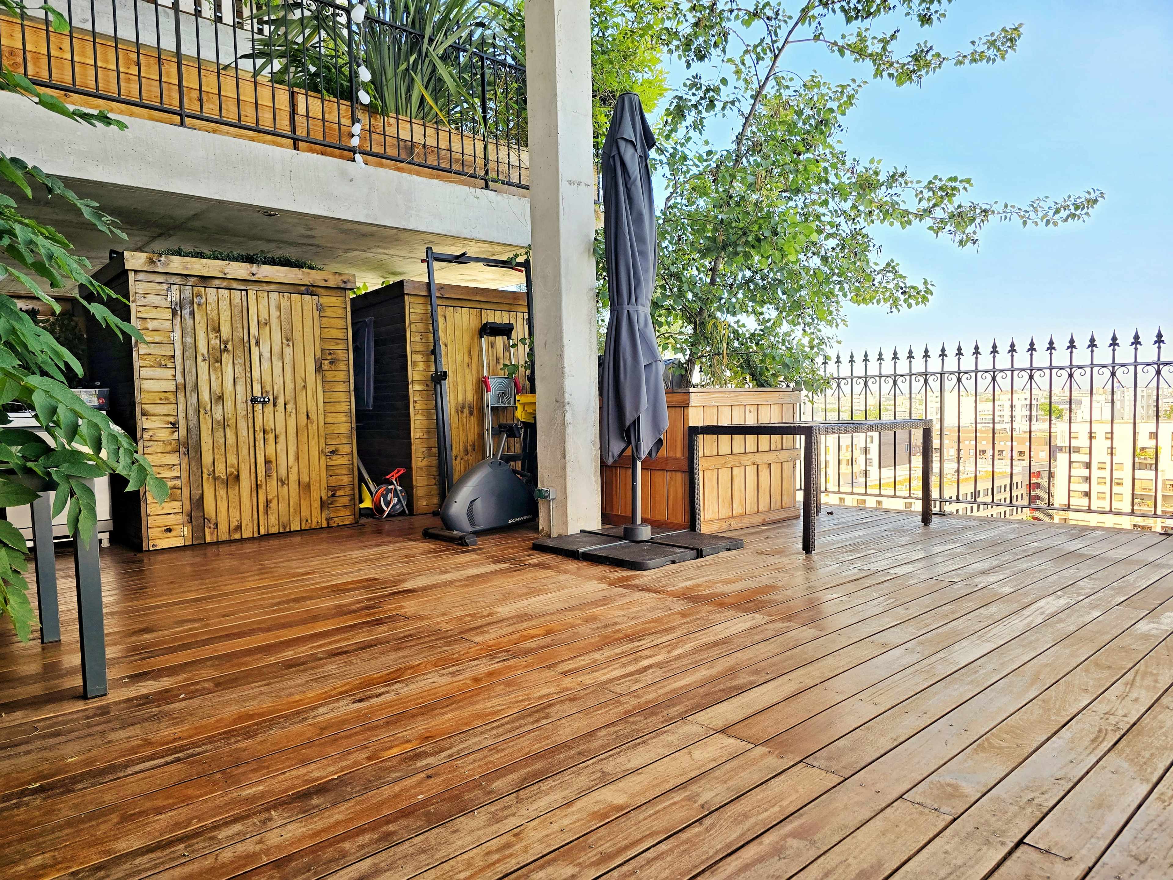 The image shows a wooden deck area with a couple of storage sheds, a patio umbrella, and greenery visible in the background.