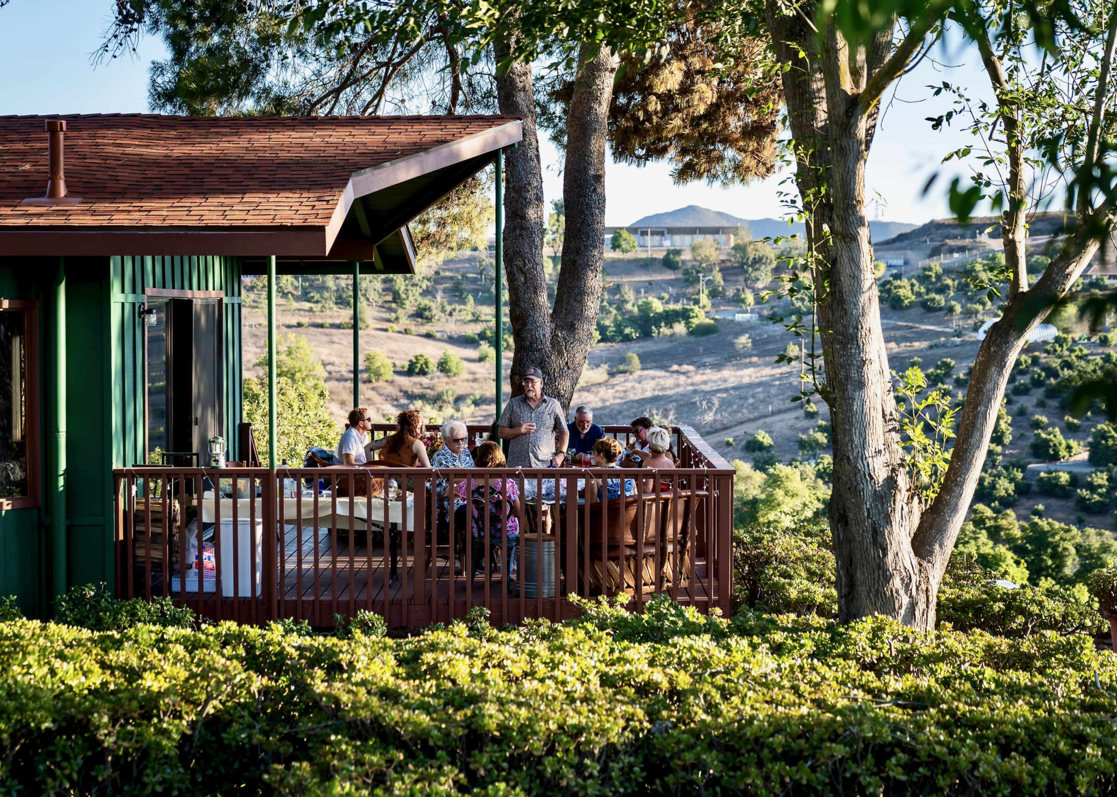 A group of people gathers on a deck outside a green house, surrounded by trees and hills.