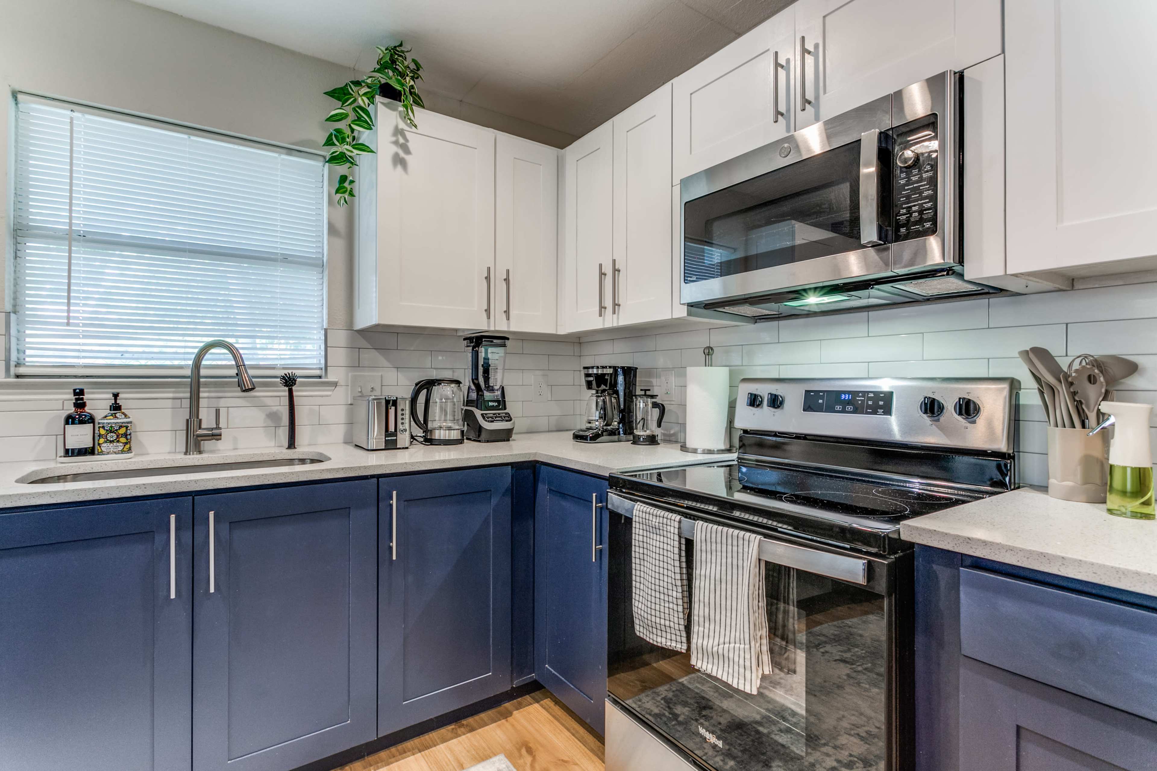 A modern kitchen features blue and white cabinetry, stainless steel appliances, and a small window with greenery.