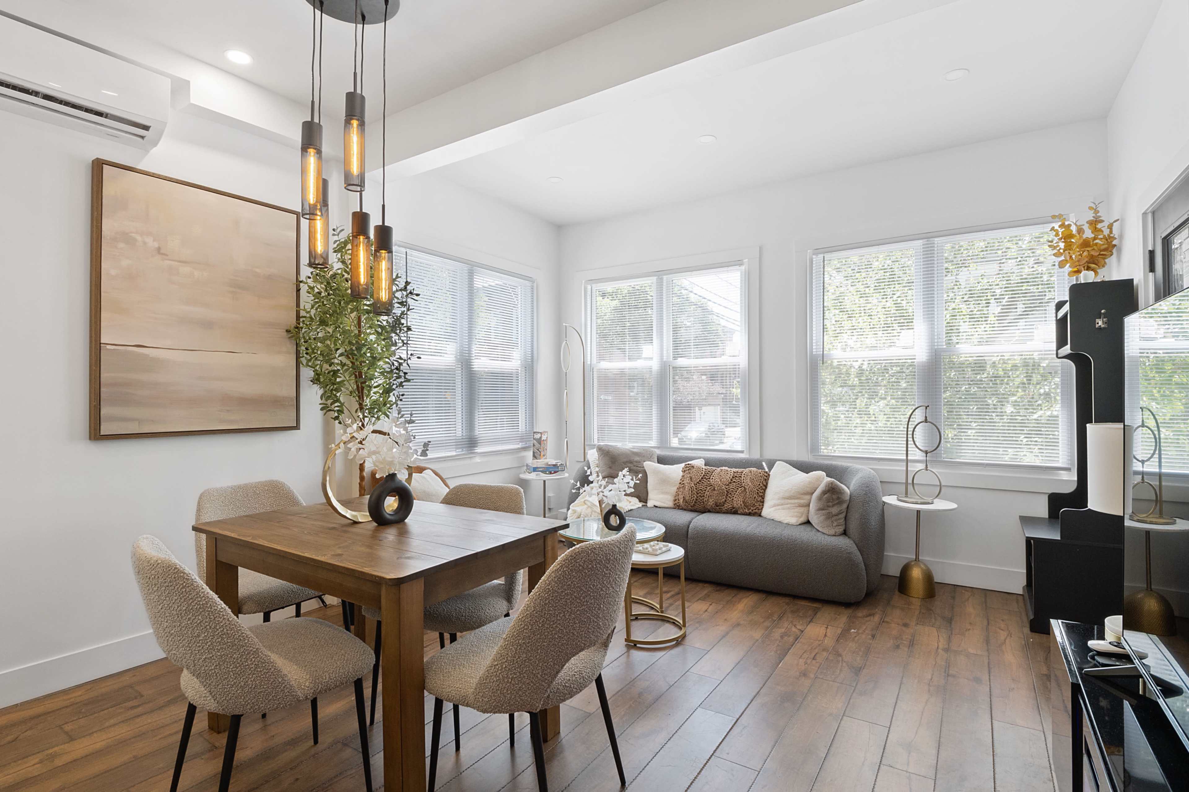 A modern living space featuring a dining table with chairs, a gray sofa, large windows, and a contemporary light fixture.