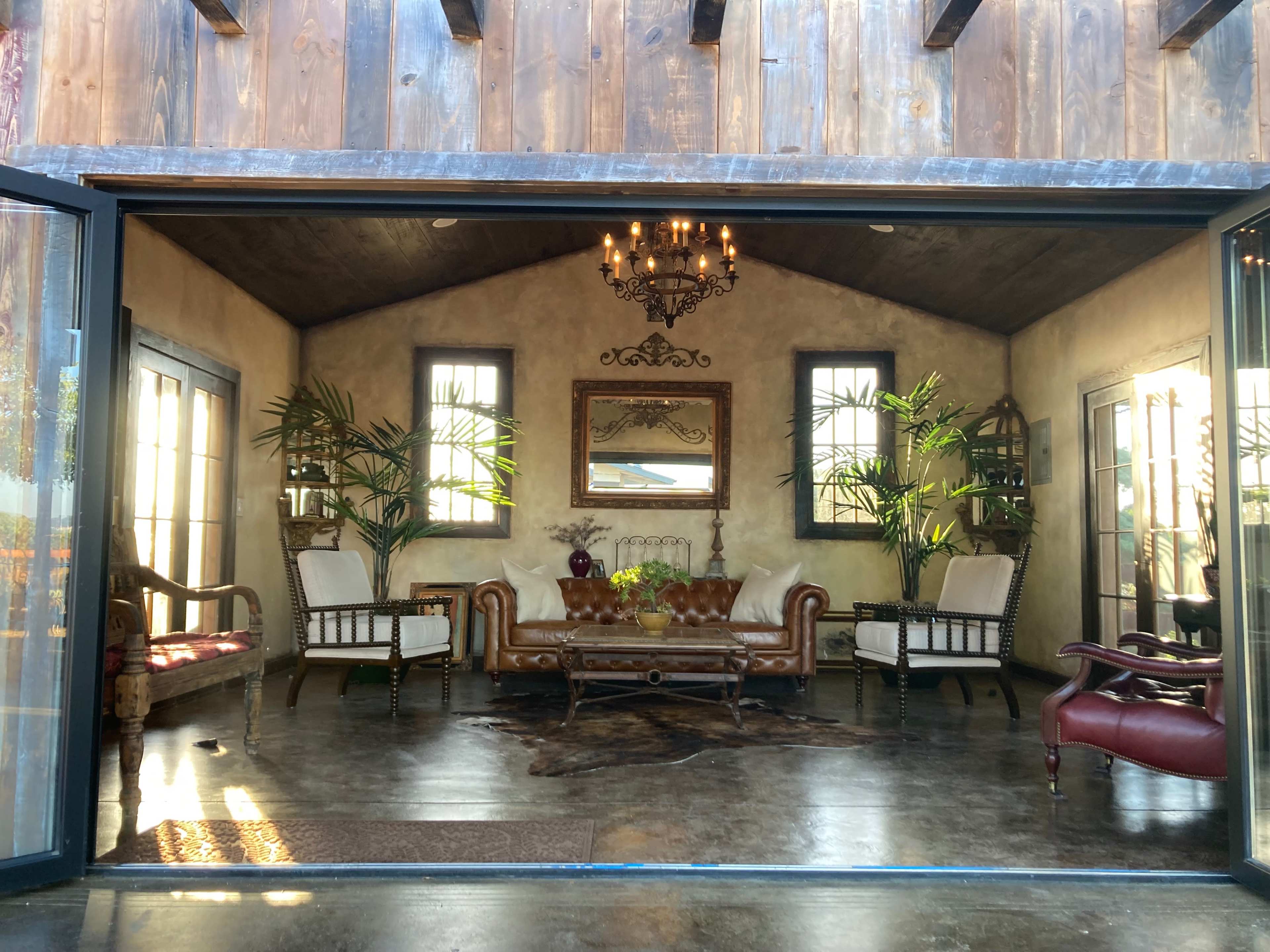 The image shows a cozy interior with a brown leather sofa, two white armchairs, a coffee table, and decorative plants under a chandelier, framed by large glass doors.