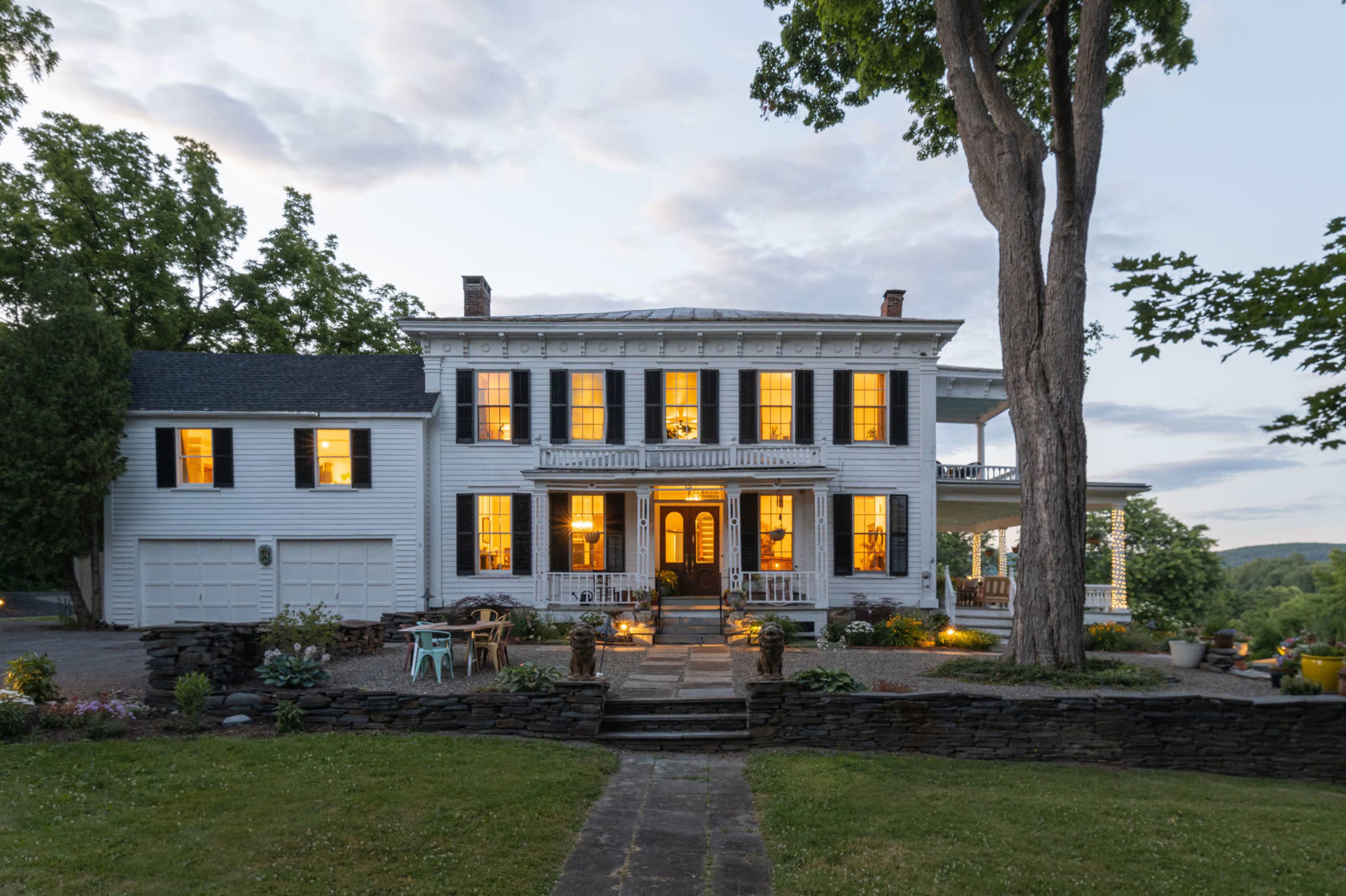 A large, two-story white house with black shutters and illuminated windows is set against a twilight sky, surrounded by trees and landscaped gardens.