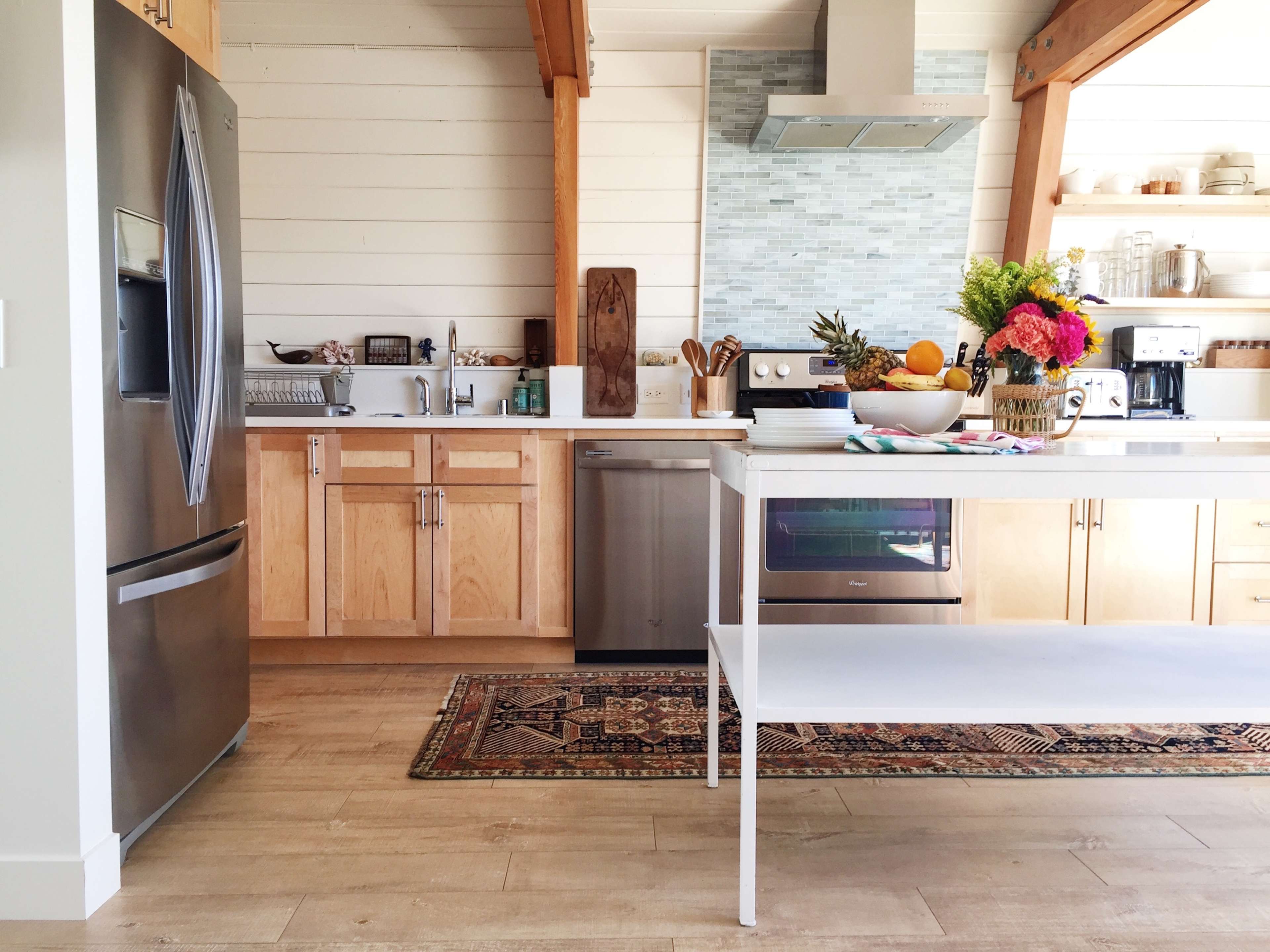 A modern kitchen features a stainless steel refrigerator, wood cabinetry, a bright countertop with a fruit arrangement, and a decorative rug on the floor.
