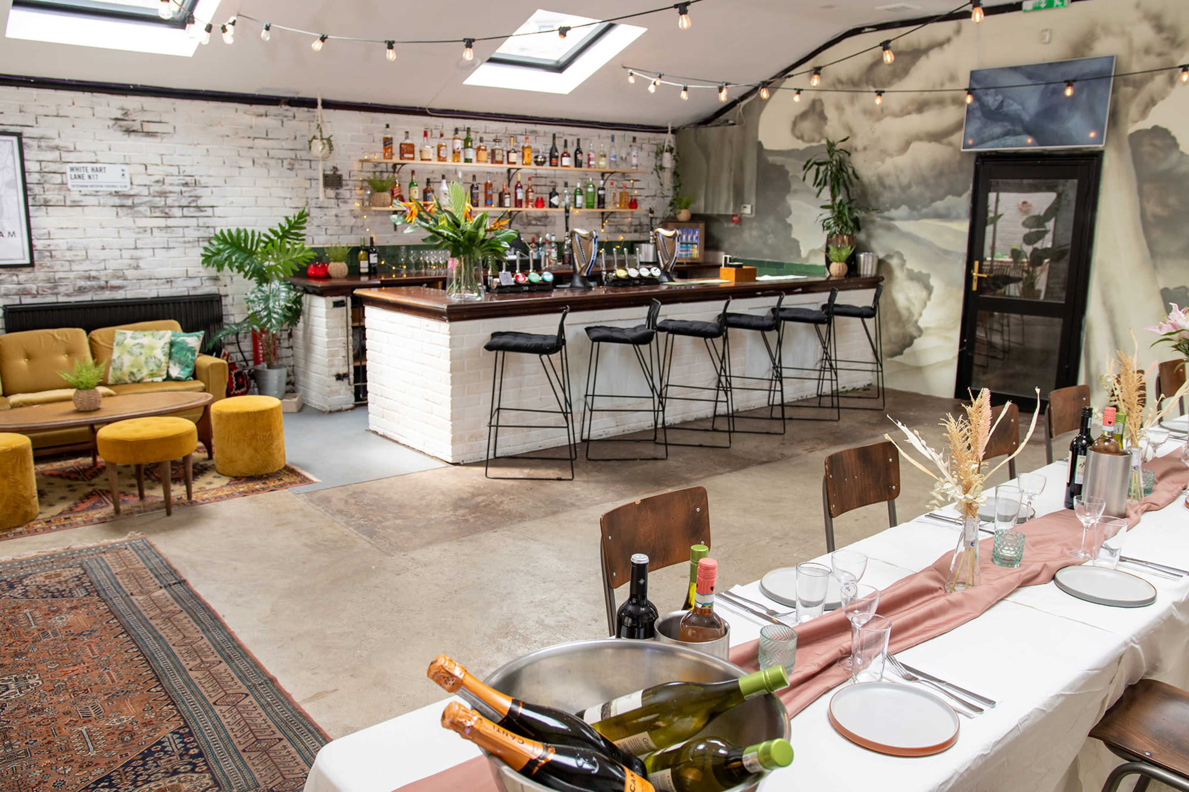A partially furnished bar area with a long table set for dining, surrounded by a variety of plants and decorative elements.