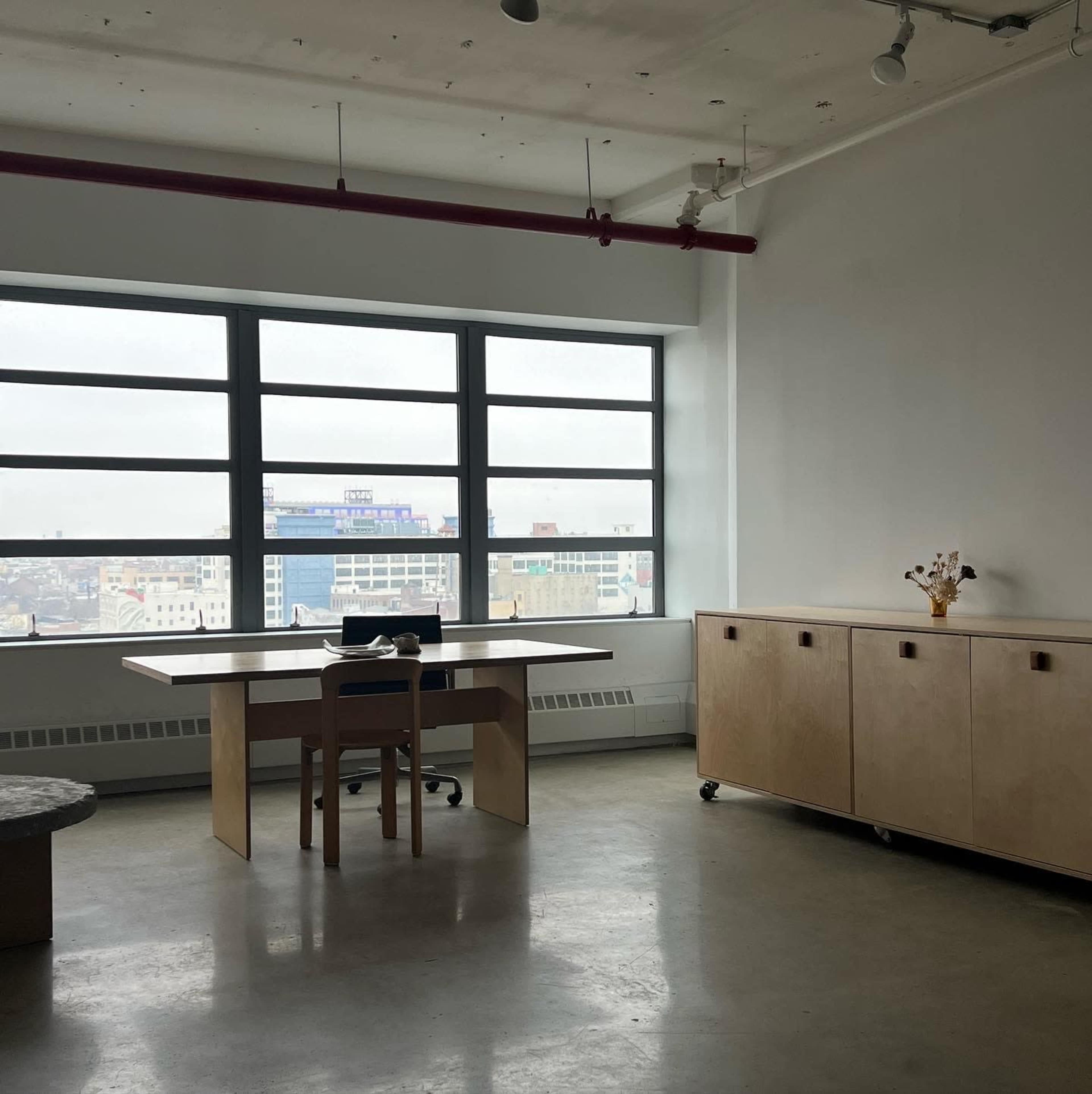 A minimalist office space features a wooden desk and cabinet, with large windows providing a view of the city skyline.
